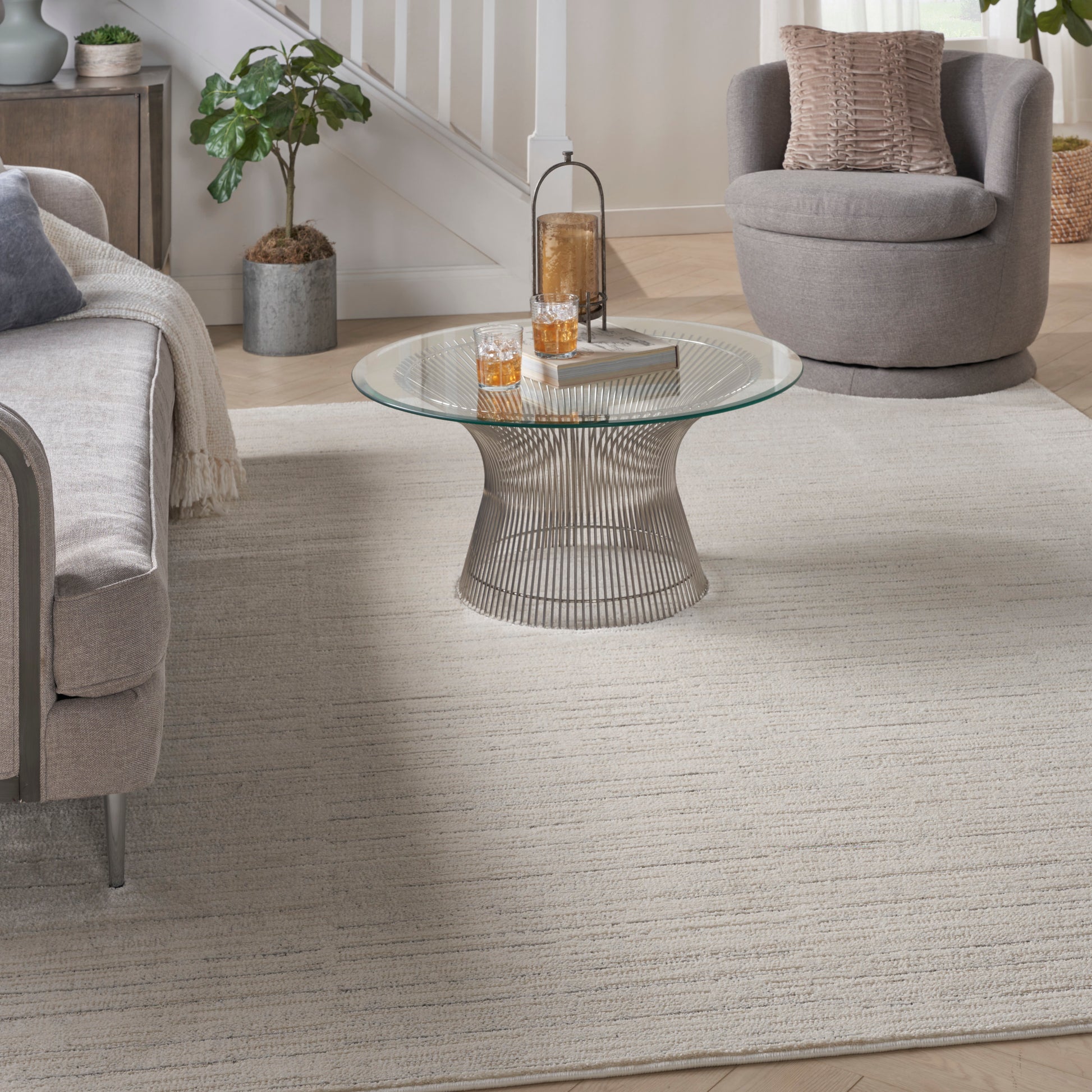 Neutral living room with round glass coffee table on a sculptural metal base, gray sofa, circular swivel chair, beige rug.