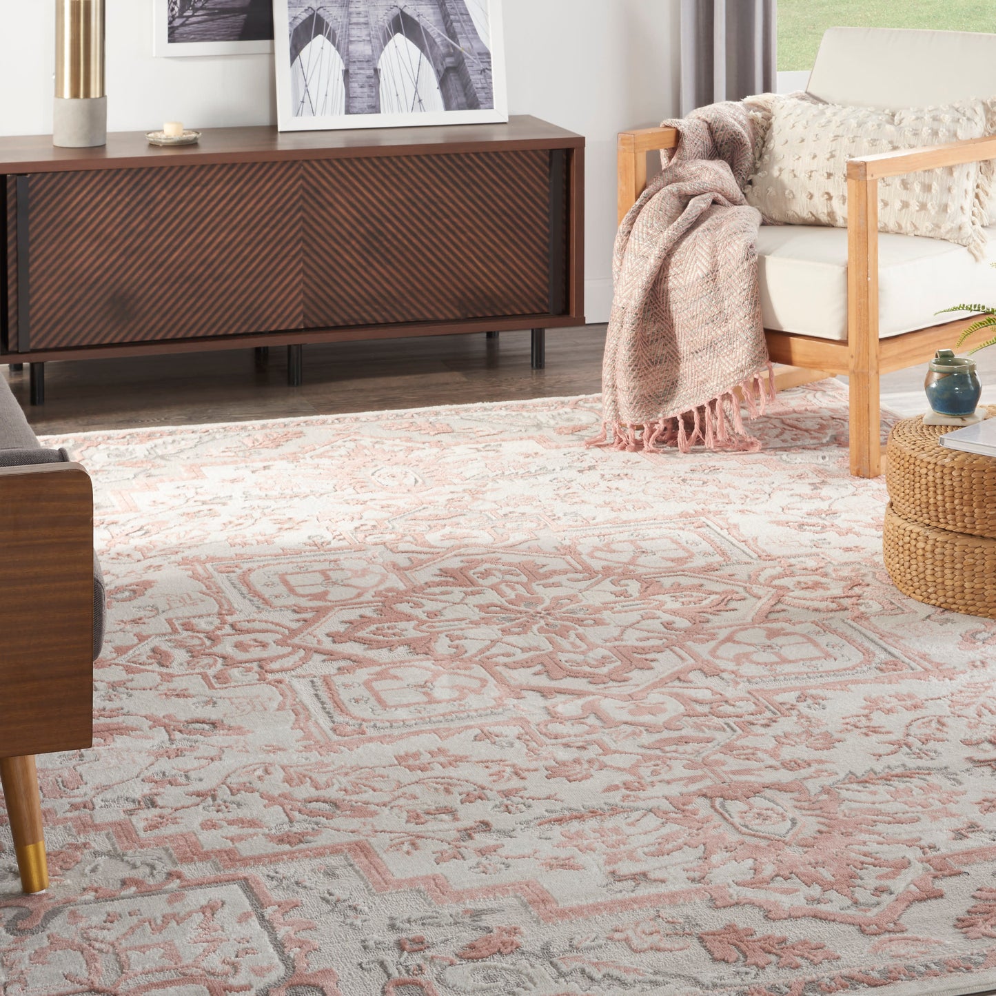 Living room with a pink patterned area rug, mid-century wood sideboard, beige sofa with knit blanket, and wicker ottoman.