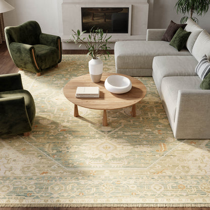 Cozy living room with round wooden coffee table, beige sectional sofa, green velvet chairs, patterned rug, and plants.