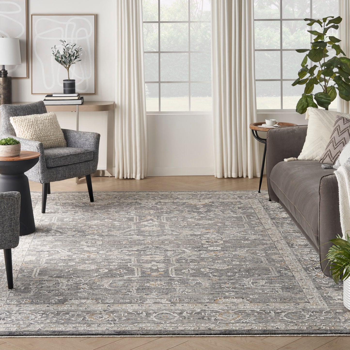 Modern living room with a distressed gray patterned area rug, gray upholstered chairs, dark sofa, cream curtains, and potted plants.