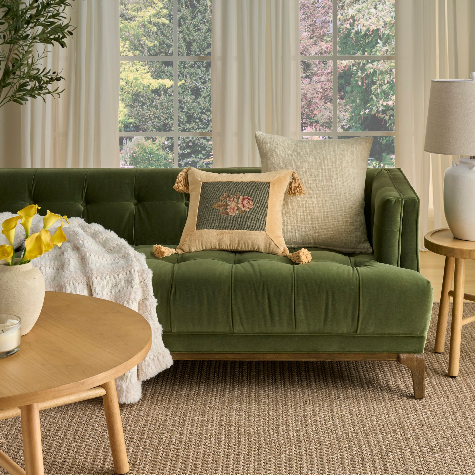 Olive green tufted velvet sofa with beige tassel cushion and cream throw, round wood coffee table with yellow flowers.