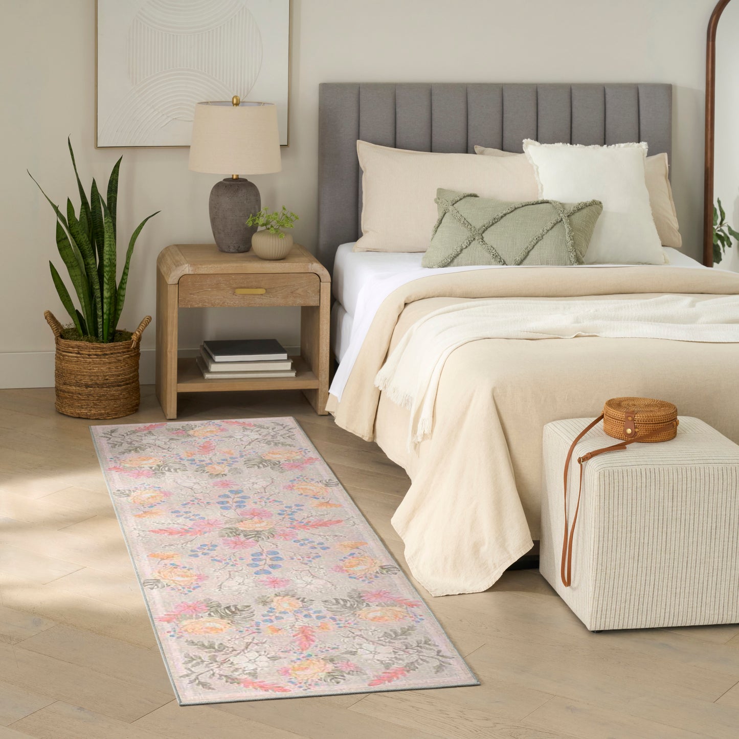 Neutral bedroom with cream bedding, gray headboard, pastel floral rug, wooden nightstand, lamp, potted plant, ottoman.