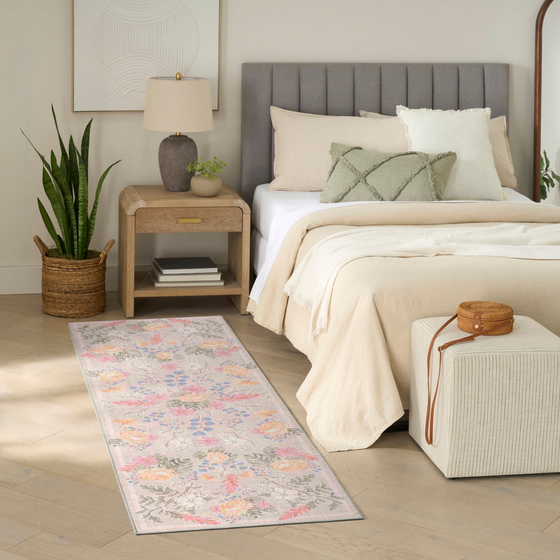 Neutral bedroom with cream bedding, gray headboard, pastel floral rug, wooden nightstand, lamp, potted plant, ottoman.