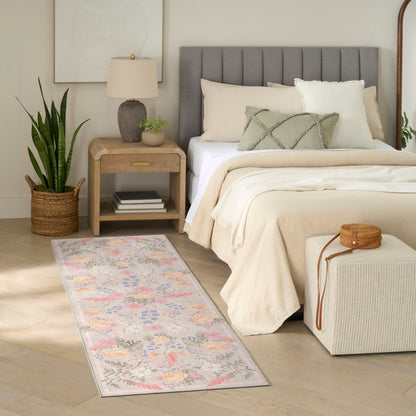 Neutral bedroom with cream bedding, gray headboard, pastel floral rug, wooden nightstand, lamp, potted plant, ottoman.