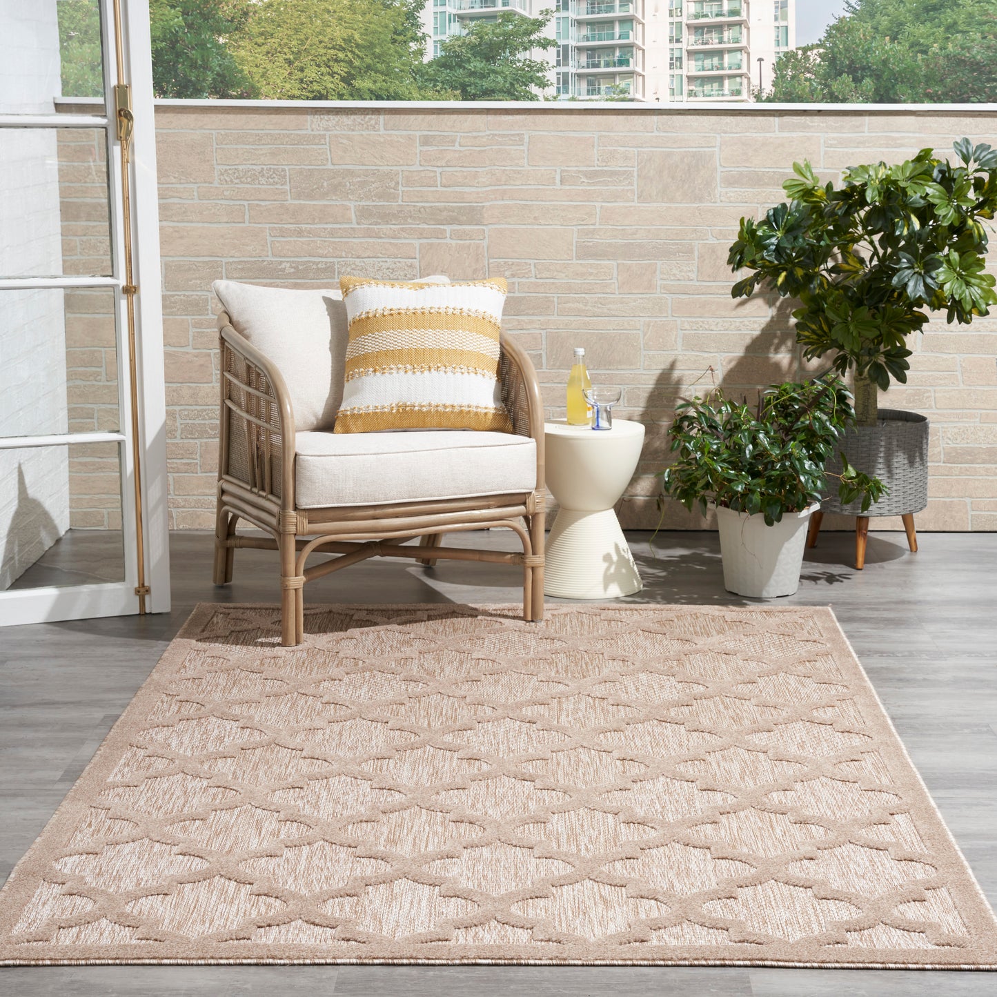 Modern 5' x 7' tan geometric rug, with wicker outdoor armchair, beige cushions, a yellow striped pillow, side table & potted plants.