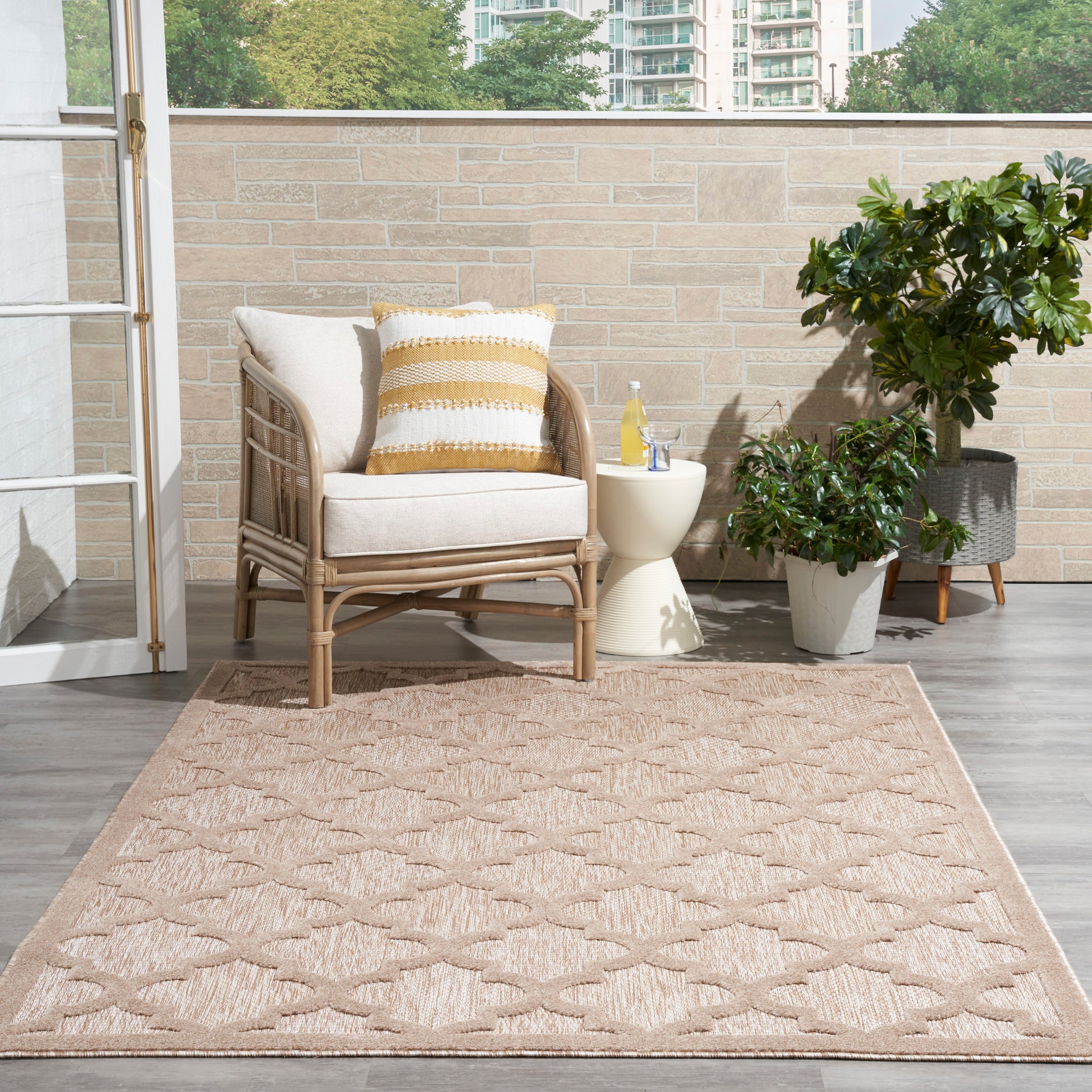 Modern 5' x 7' tan geometric rug, with wicker outdoor armchair, beige cushions, a yellow striped pillow, side table & potted plants.
