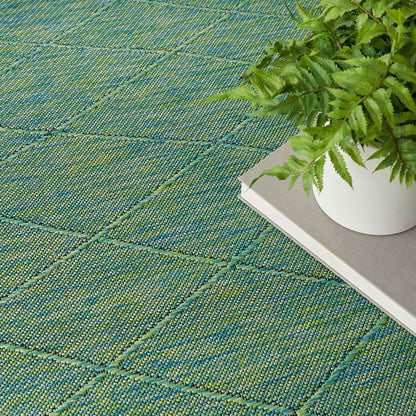 Green woven rug with teal grid lines, white ceramic pot with a fern on a gray-covered book.