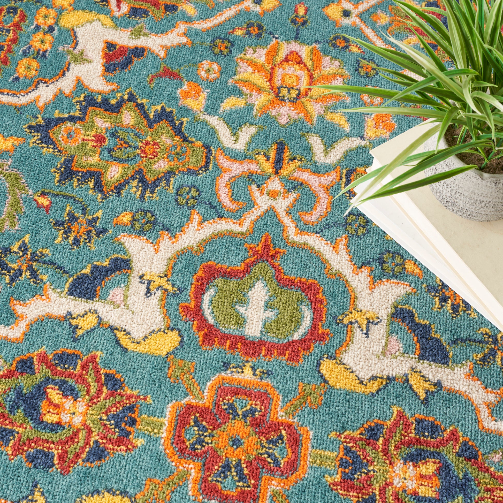 Turquoise Bohemian round rug with orange, red, yellow and cream floral motifs; woven pile, shown beside a potted plant and books.