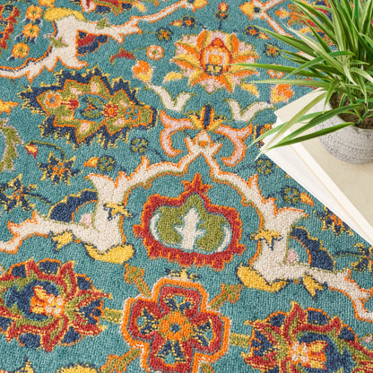Turquoise Bohemian round rug with orange, red, yellow and cream floral motifs; woven pile, shown beside a potted plant and books.