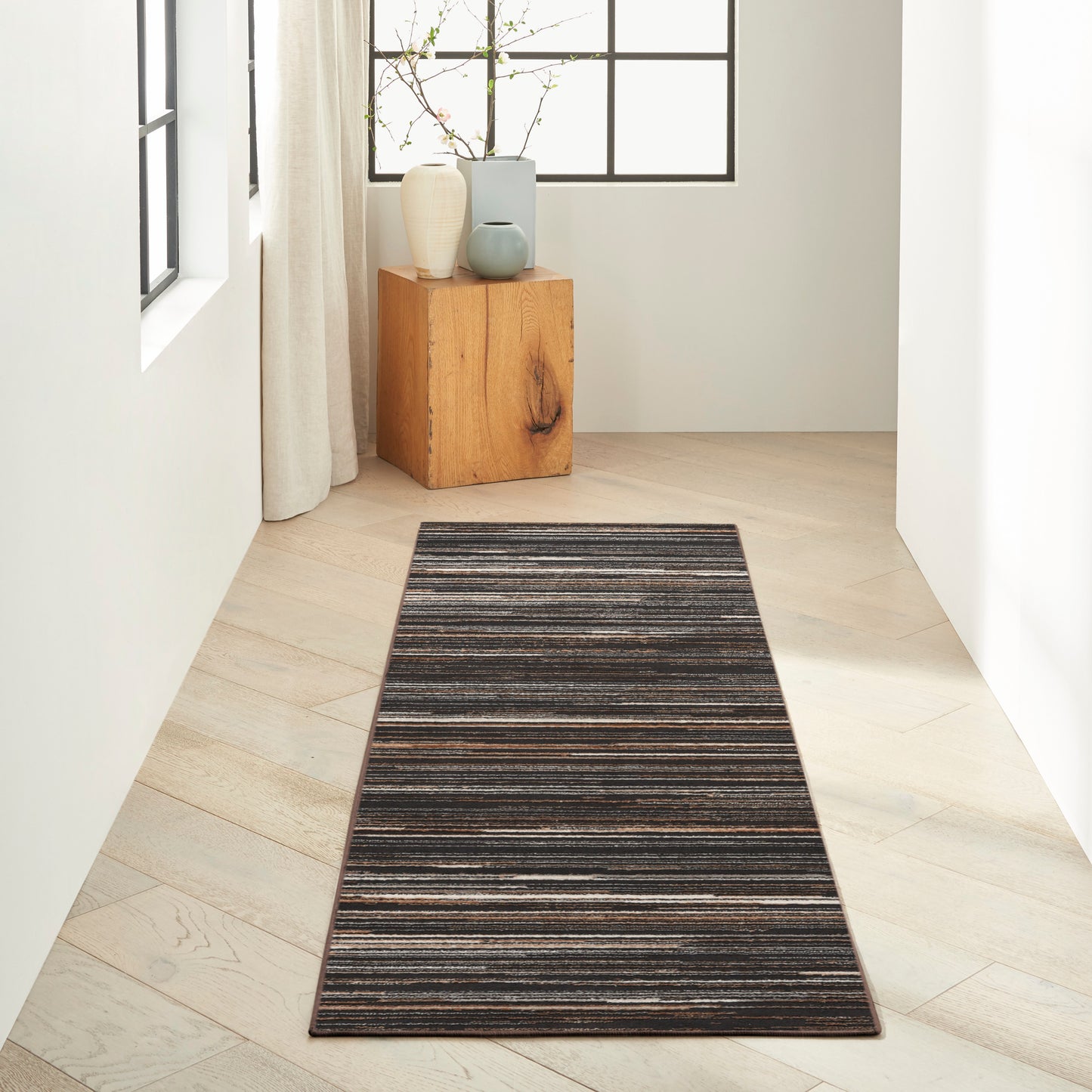 Narrow hallway with a dark striped runner rug on pale wood flooring, a wooden cube table with vases by a window.