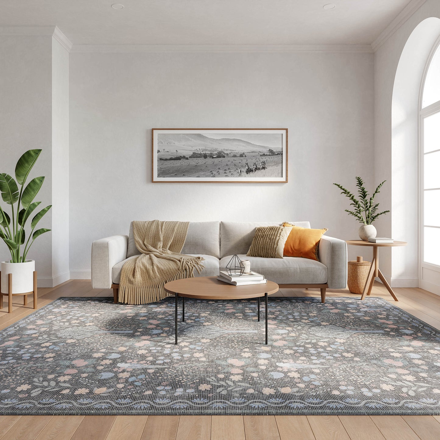 Light living room with gray sofa, beige throw, round wood coffee table, blue-gray rug, plants, and arched window.