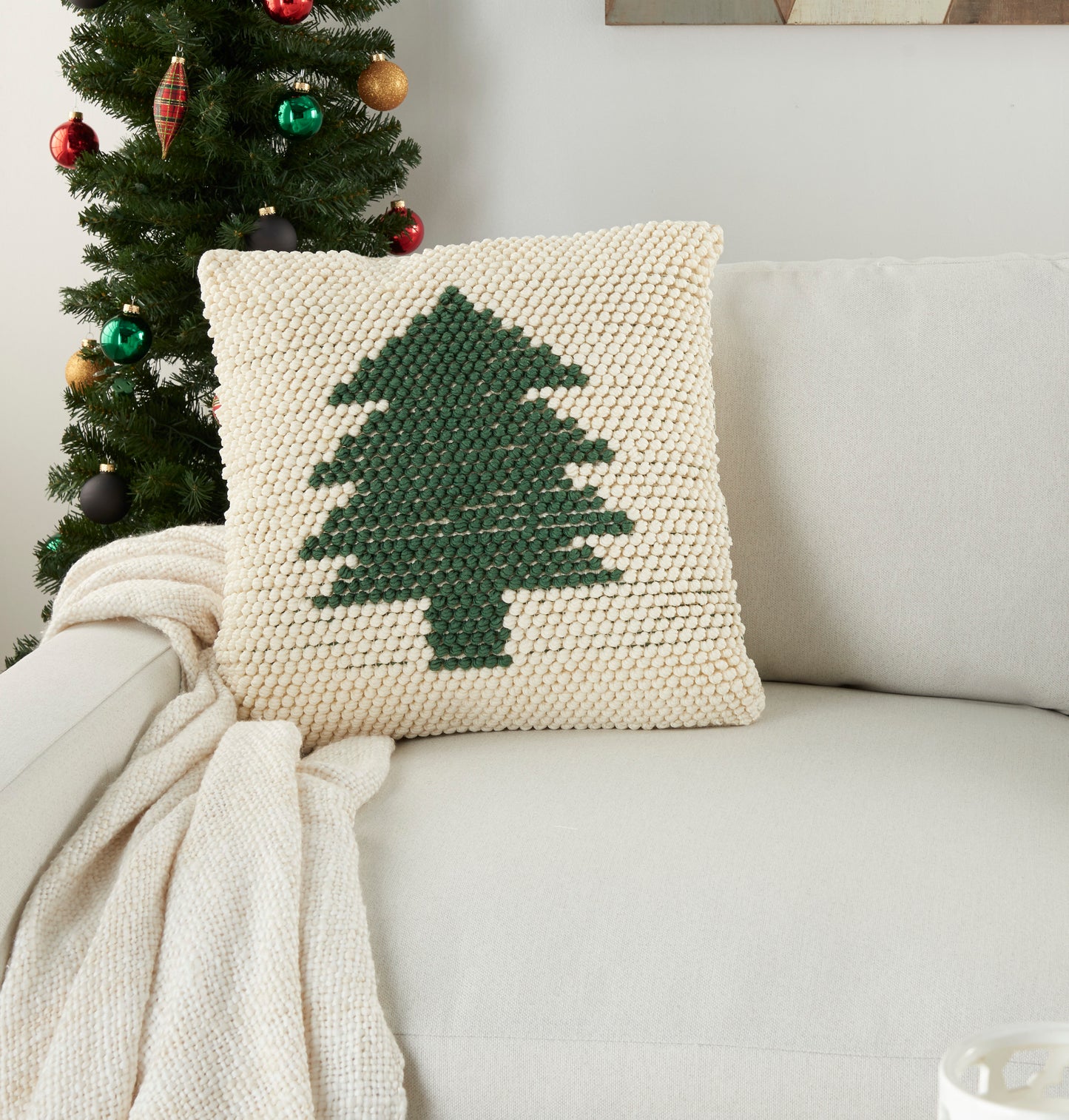 Cream chunky-knit square pillow with dark green Christmas tree motif on a beige sofa, paired with a cozy knit throw.