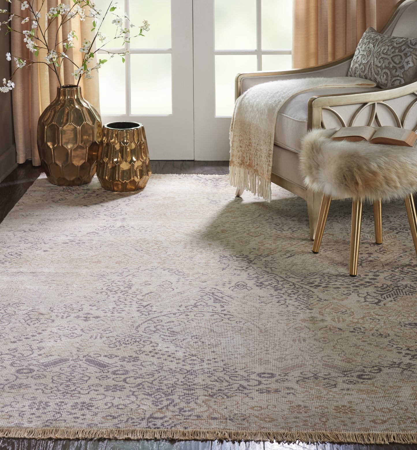 Soft beige patterned area rug with fringe, gold hammered-metal vases, a fur stool, and a cream sofa in a sunlit living room.