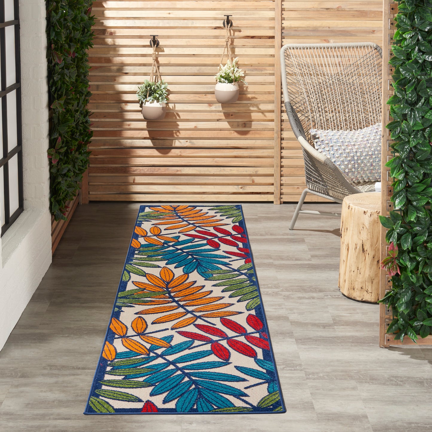 Colorful outdoor runner rug with blue, orange, red, green leaf pattern on a wooden patio with wicker chair and plants.