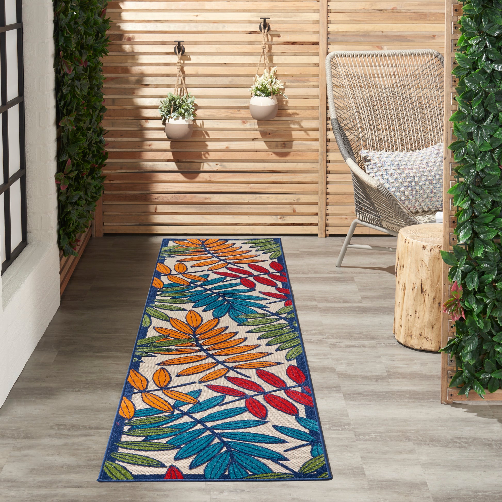 Colorful outdoor runner rug with blue, orange, red, green leaf pattern on a wooden patio with wicker chair and plants.