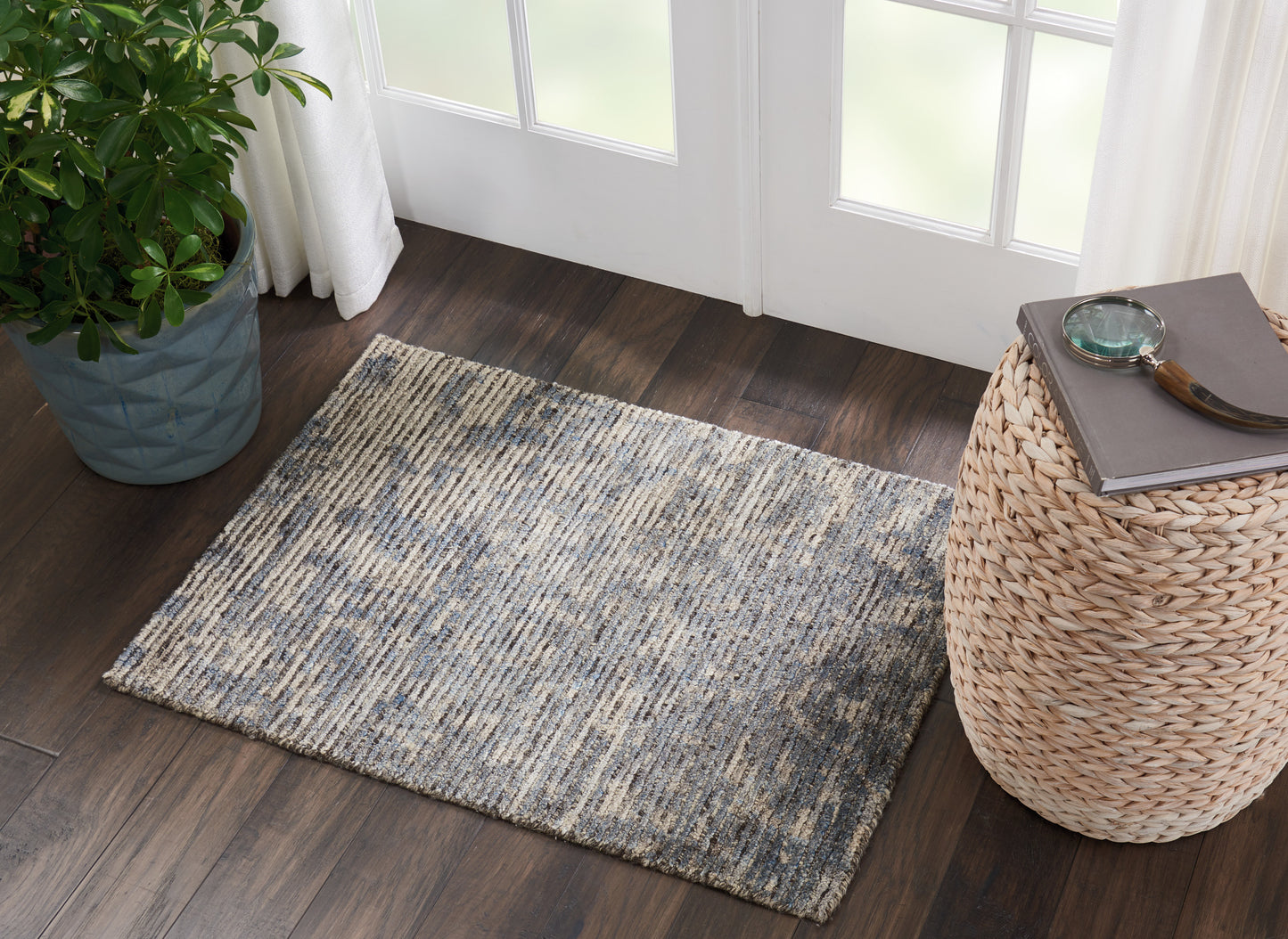 Rectangular blue-gray rug on dark wood floor beside a woven wicker side table with a book and magnifier, near a potted plant.