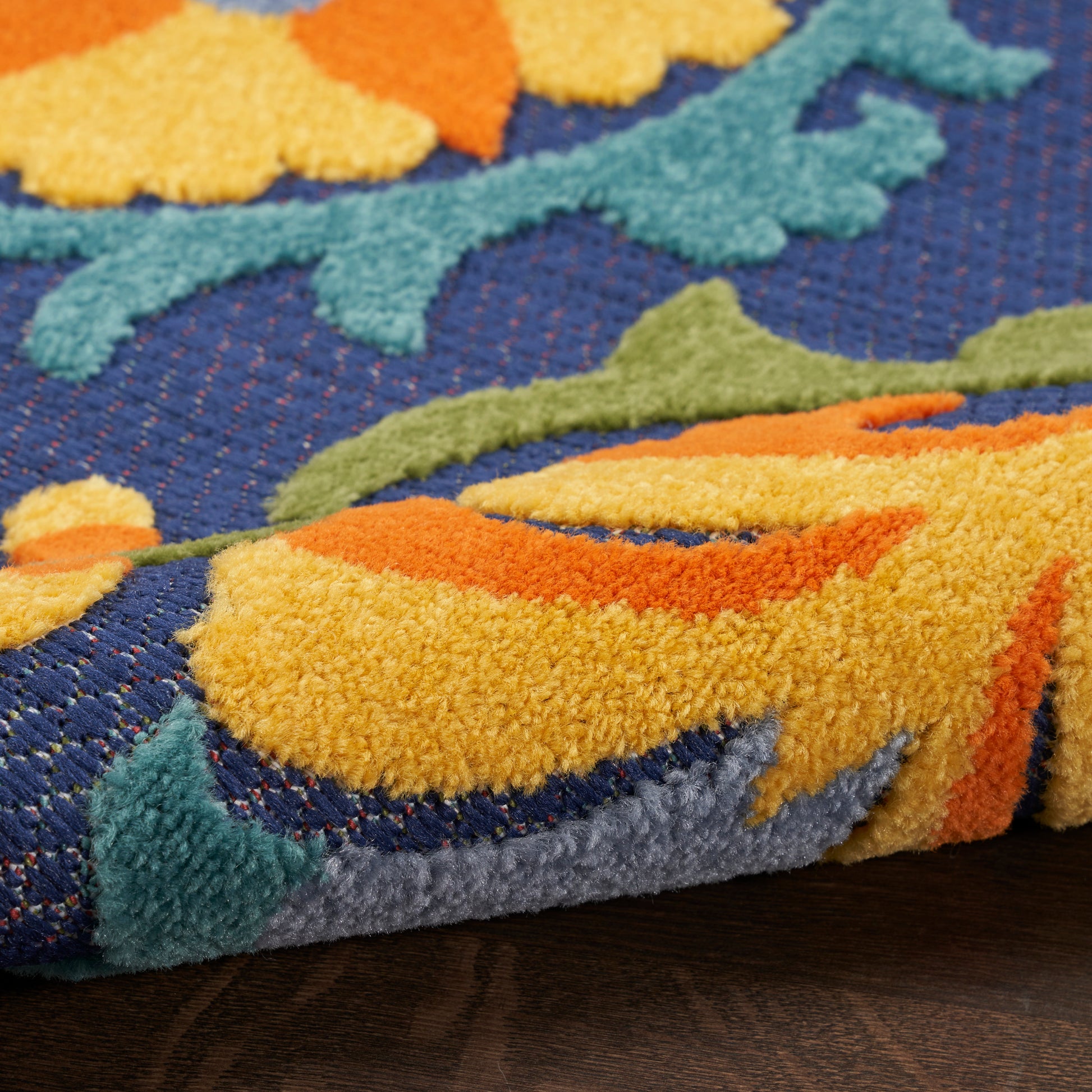 Close-up of vibrant tufted rug with navy base and yellow, orange, teal floral pattern, thick pile on wood floor.