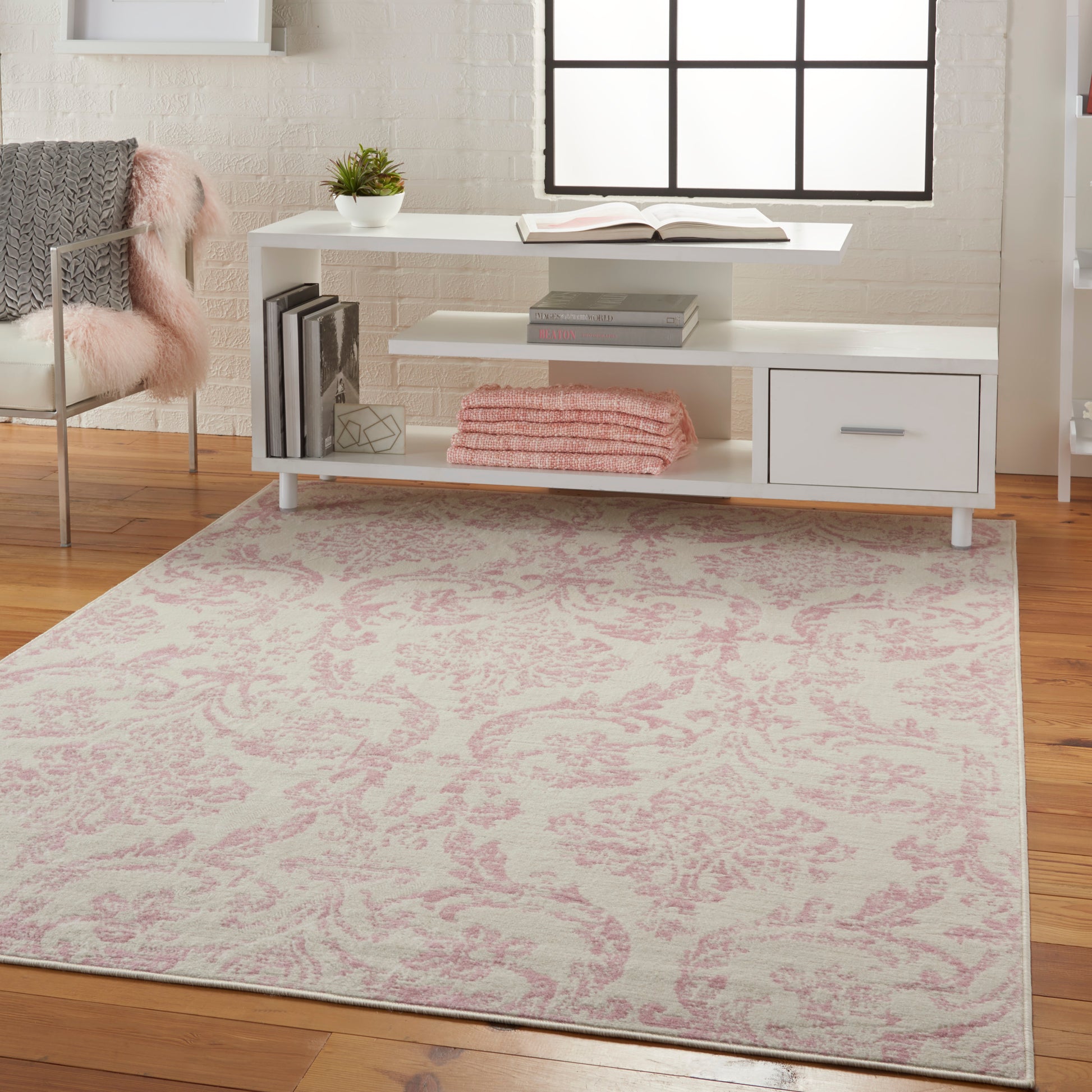 White low-profile console with shelves and drawer, pink damask rug, pink towels, plant, book, and a chair with pink faux fur.