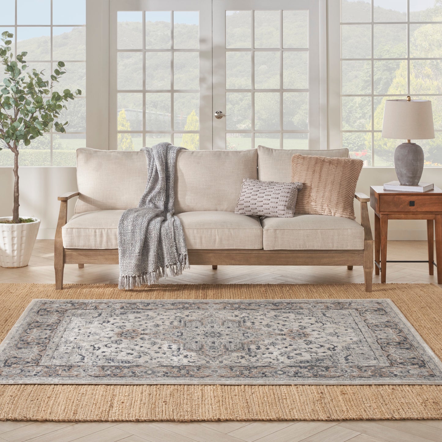 Cream sofa with wooden legs, gray throw and cushions, on a patterned rug over jute, with a side table and lamp.