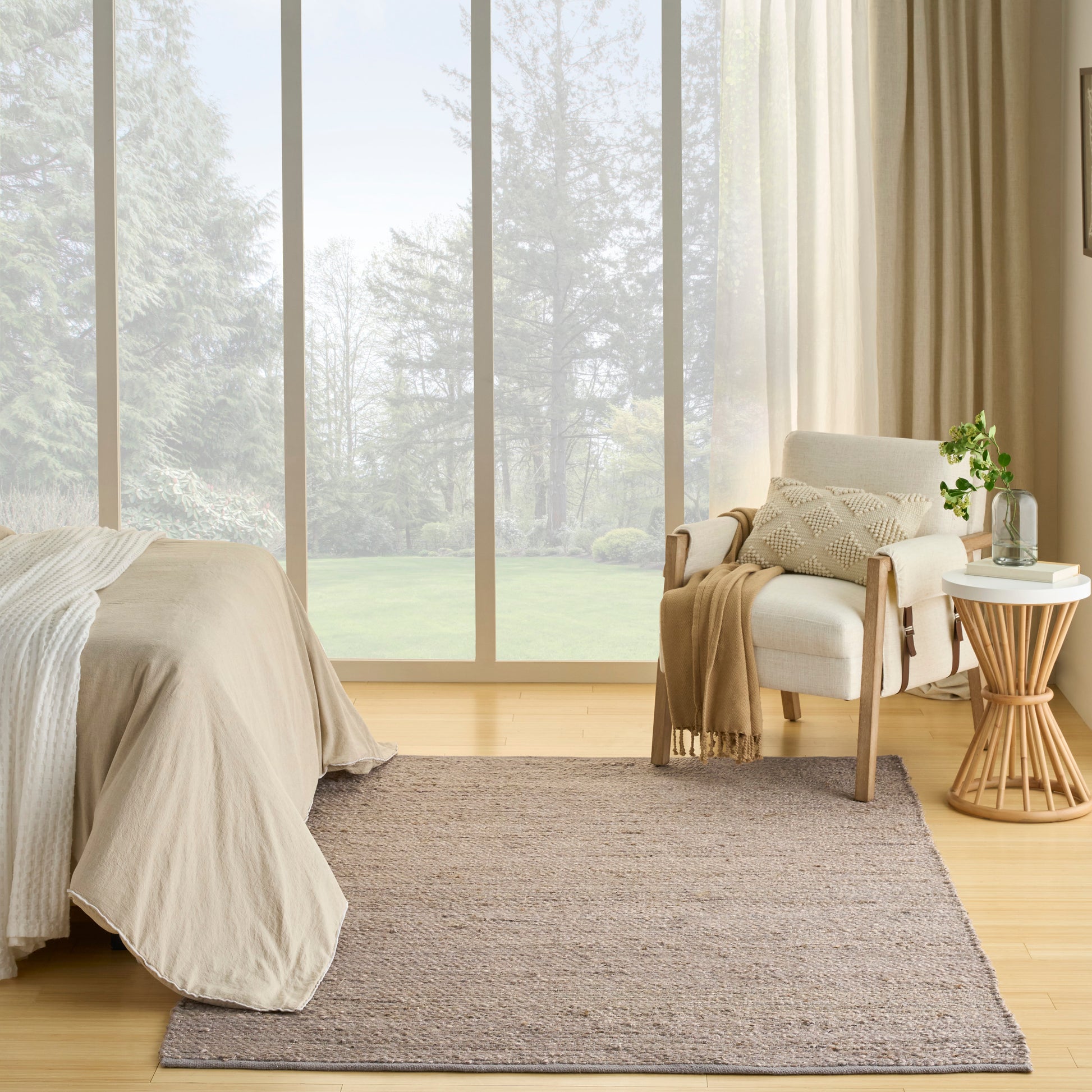 Neutral bedroom with large glass wall, beige bedding, woven rug, and an armchair with a round wooden side table and plant.