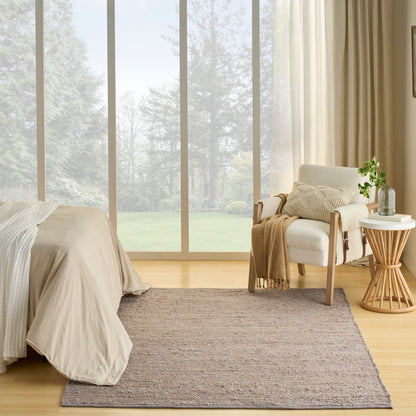 Neutral bedroom with large glass wall, beige bedding, woven rug, and an armchair with a round wooden side table and plant.