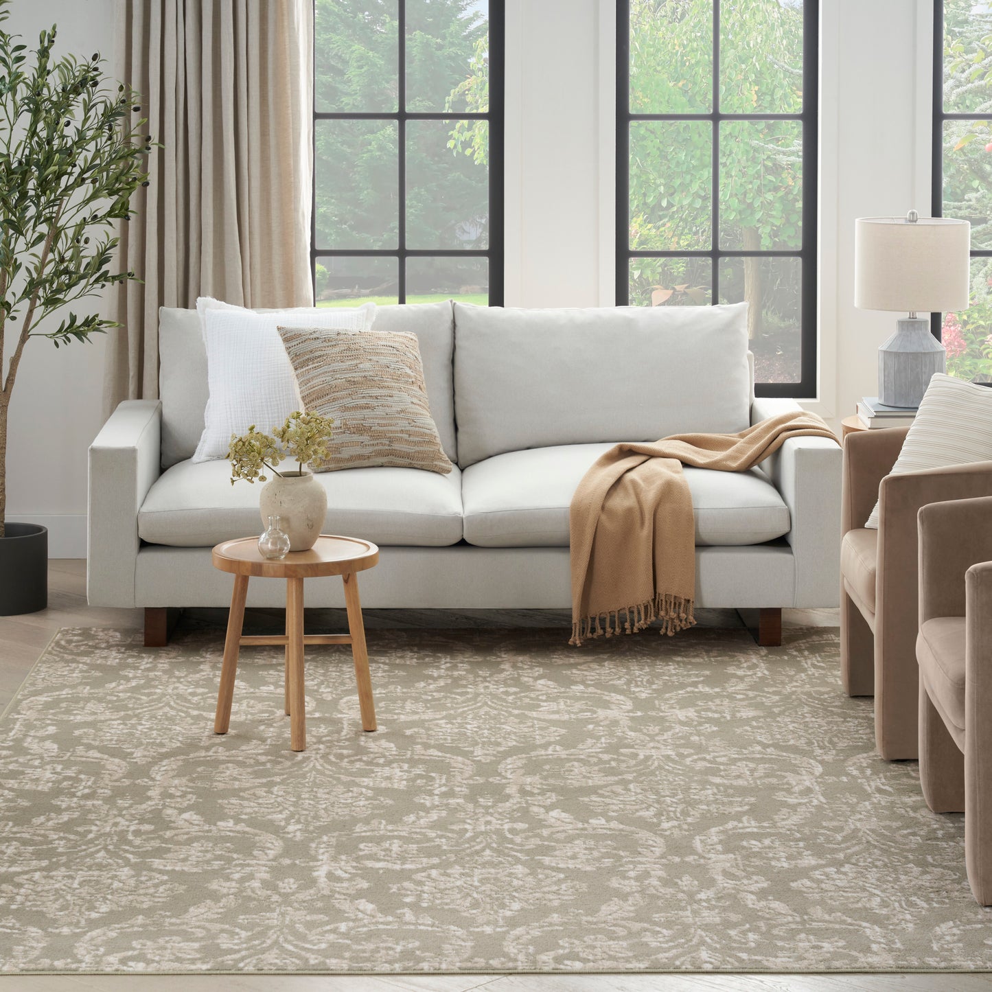 Bright living room with light gray sofa, beige throw, damask rug, round wood side table, and neutral beige chairs.