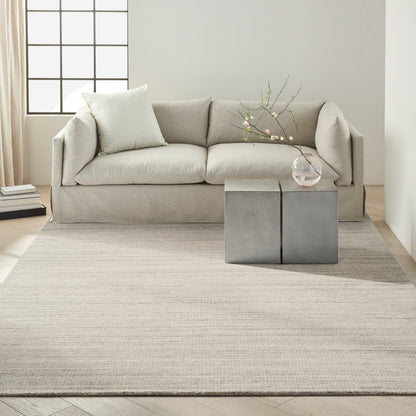 Beige fabric sofa with light cushions, grey cube side tables and a glass vase on a pale rug in a minimalist living room.