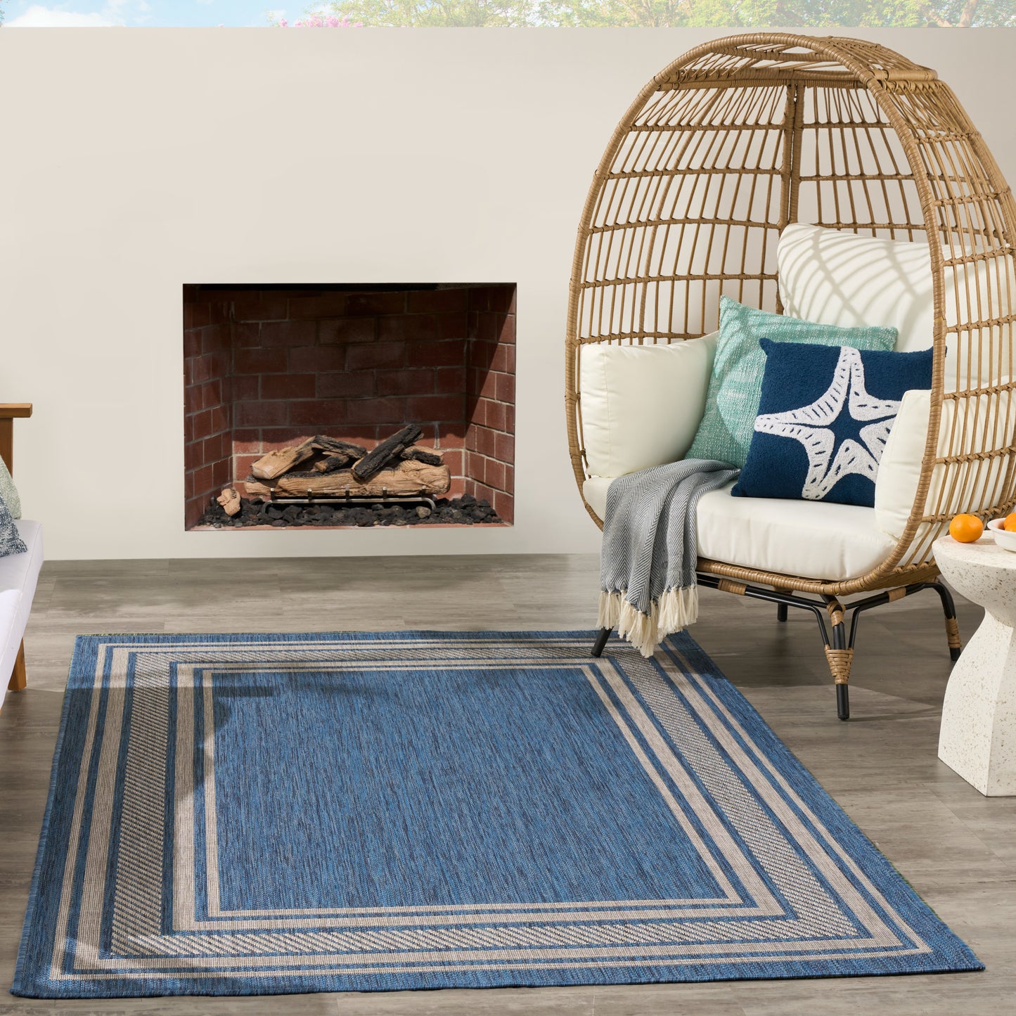 Blue striped outdoor rug on wooden deck, wicker egg chair with white cushions and starfish pillow, near brick fireplace.