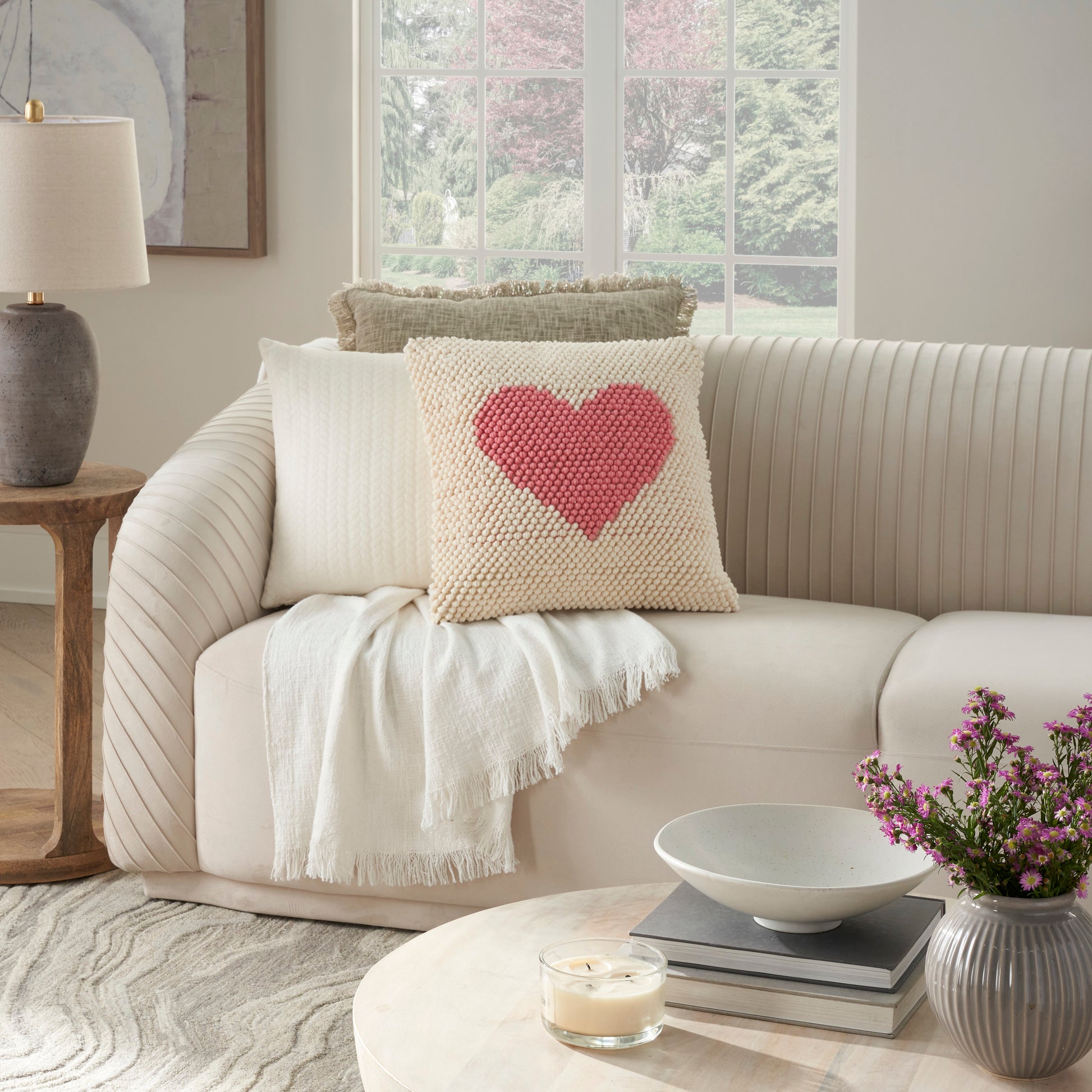 Beige knitted cushion with pink heart on cream sofa, white throw blanket; cozy modern living room decor.