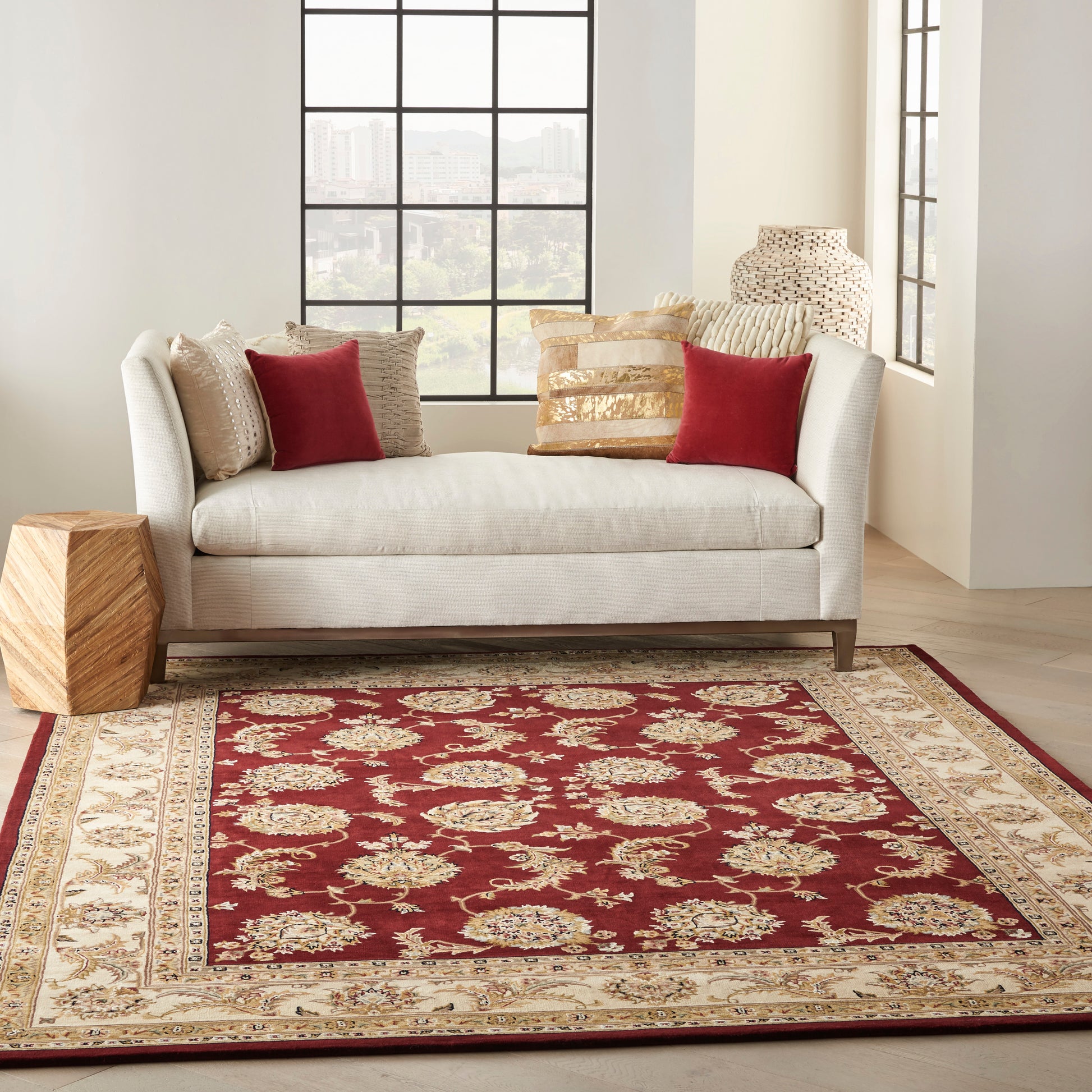 Rectangular oriental rug in red with ivory and gold border under a light gray sofa with red and gold pillows.