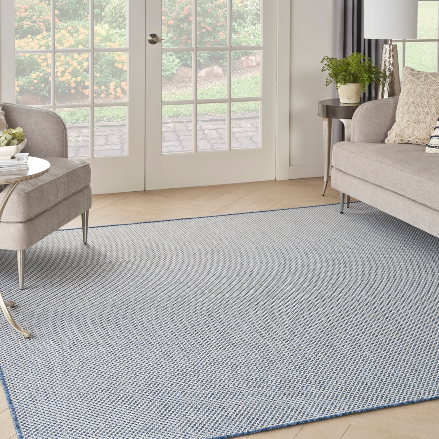 Soft blue-gray woven area rug in a beige living room with two light sofas, glass doors, and a small side table with plant.