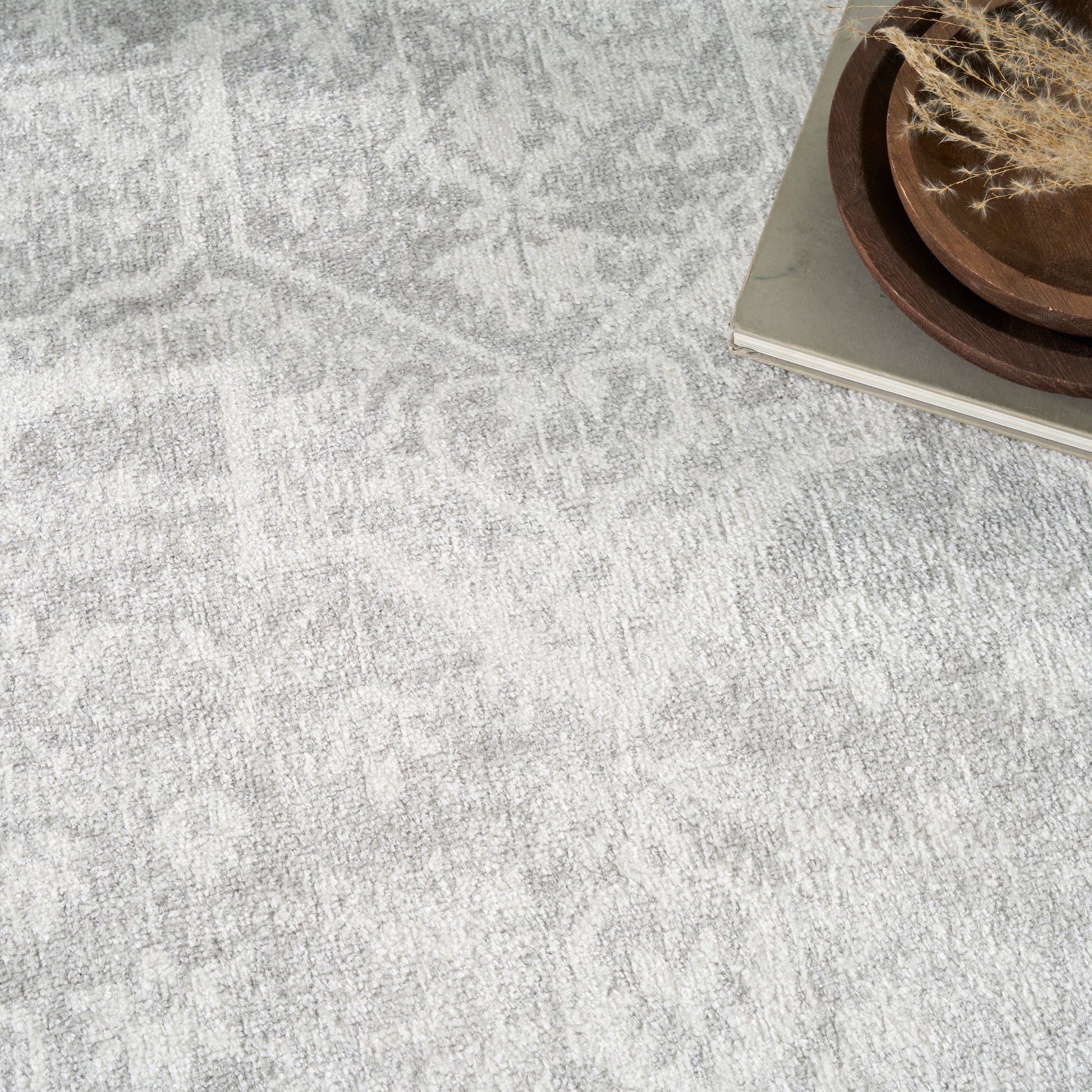 Light gray textured rug with a subtle geometric pattern, beside a stack of books and a wooden dish with dried grasses.