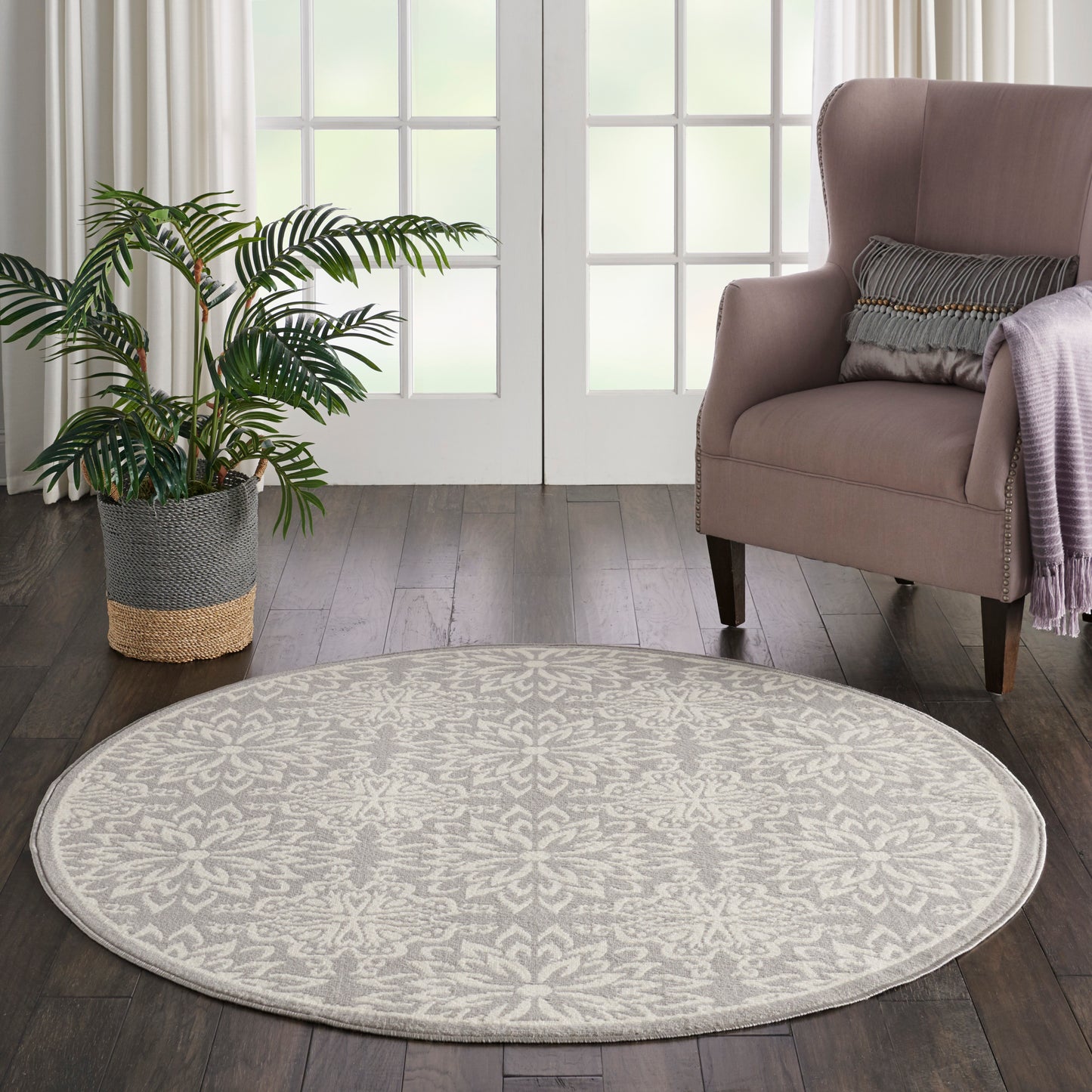 Round beige rug on dark wood floor beside pink armchair, striped cushion, lavender throw, and a potted plant by glass doors.