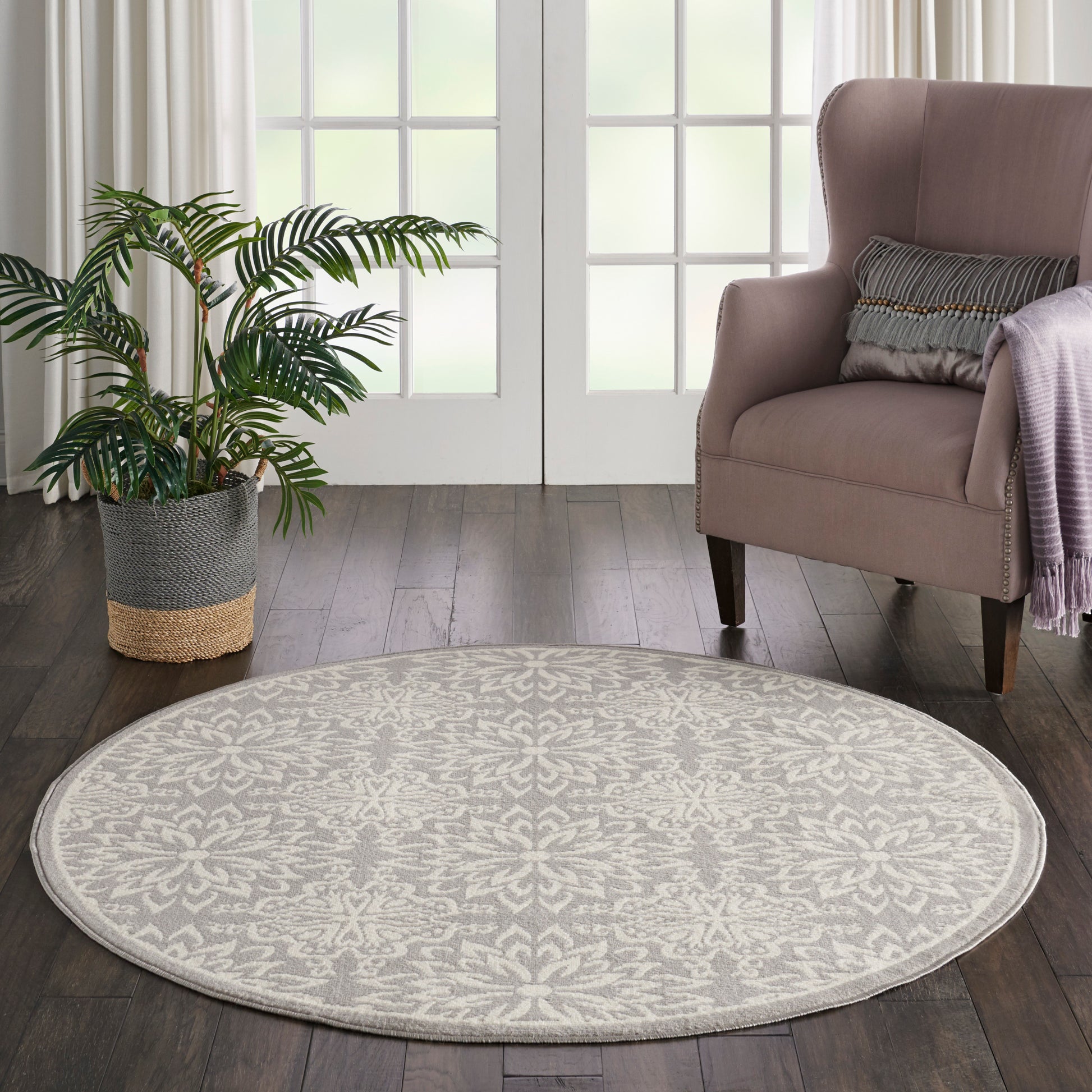 Round beige rug on dark wood floor beside pink armchair, striped cushion, lavender throw, and a potted plant by glass doors.