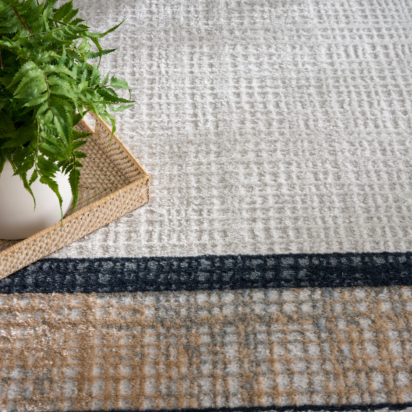 Green fern in a white pot on a woven straw tray, set on a beige textured rug with a navy stripe.