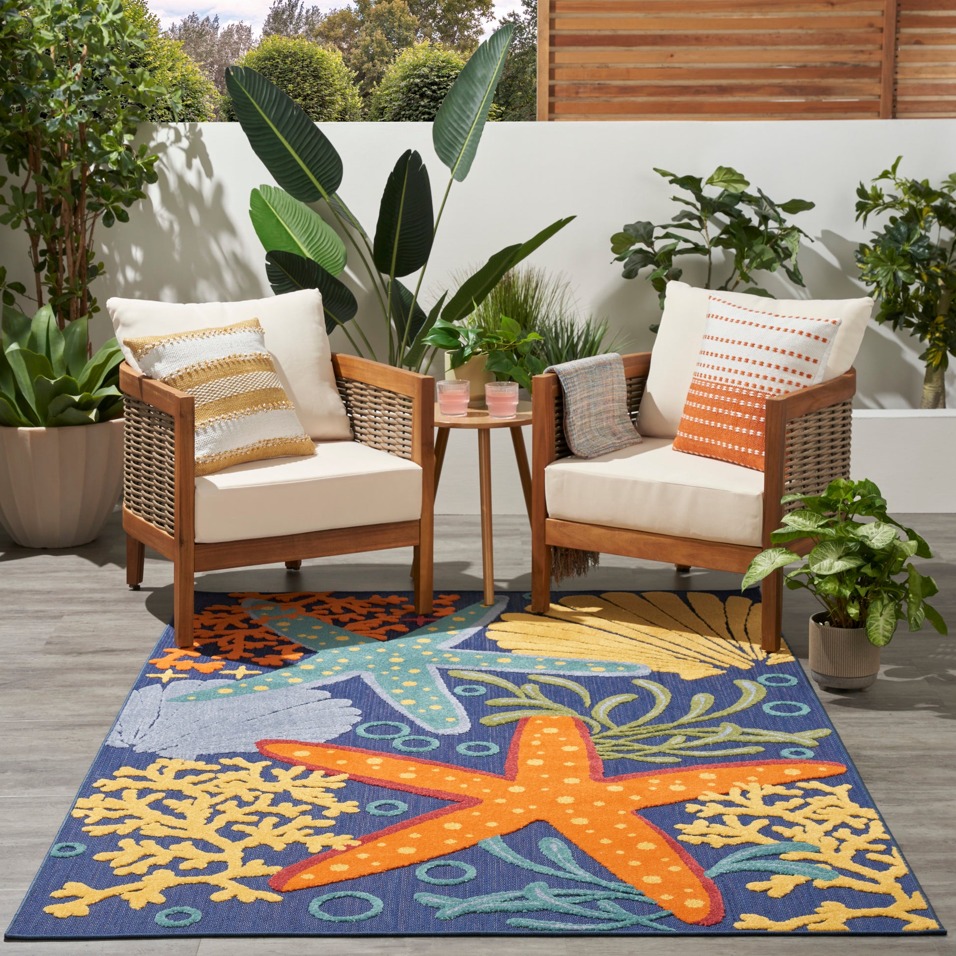 Outdoor rug 4' x 6' with a blue field and orange starfish pattern, on a patio beside wood-framed cushioned chairs, a table, and potted tropical plants.