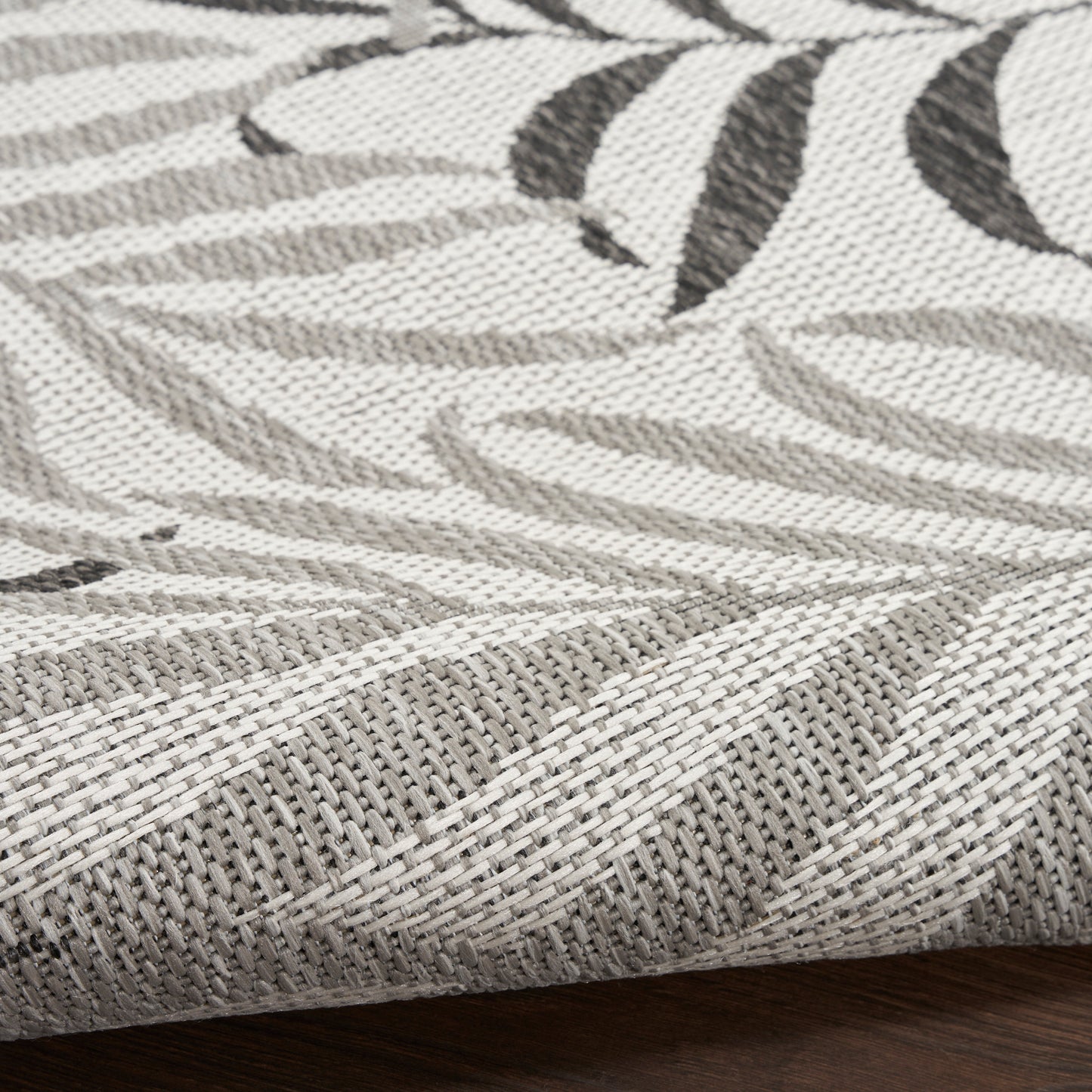 Gray-taupe woven rug with leaf pattern, textured jacquard weave, folded corner on wood floor.