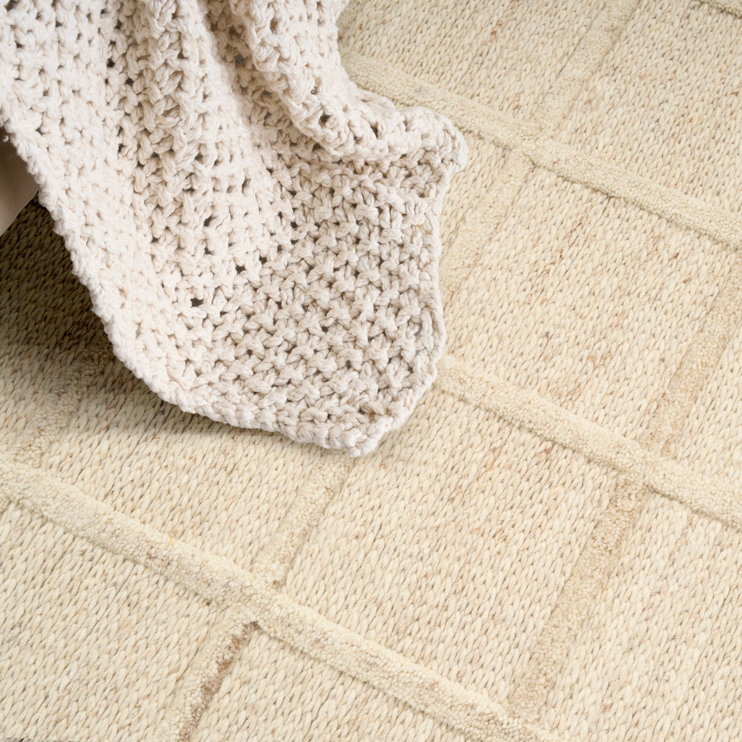 Beige textured rug with a square grid pattern, a cream chunky knit blanket draped over it.