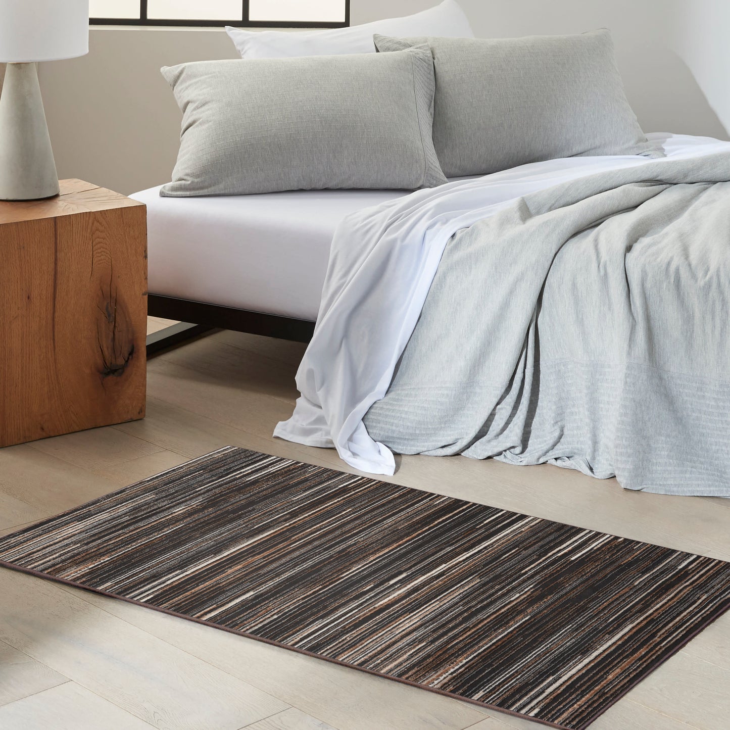 Brown-gray striped area rug beside a white bed with light gray bedding; wooden nightstand with lamp.