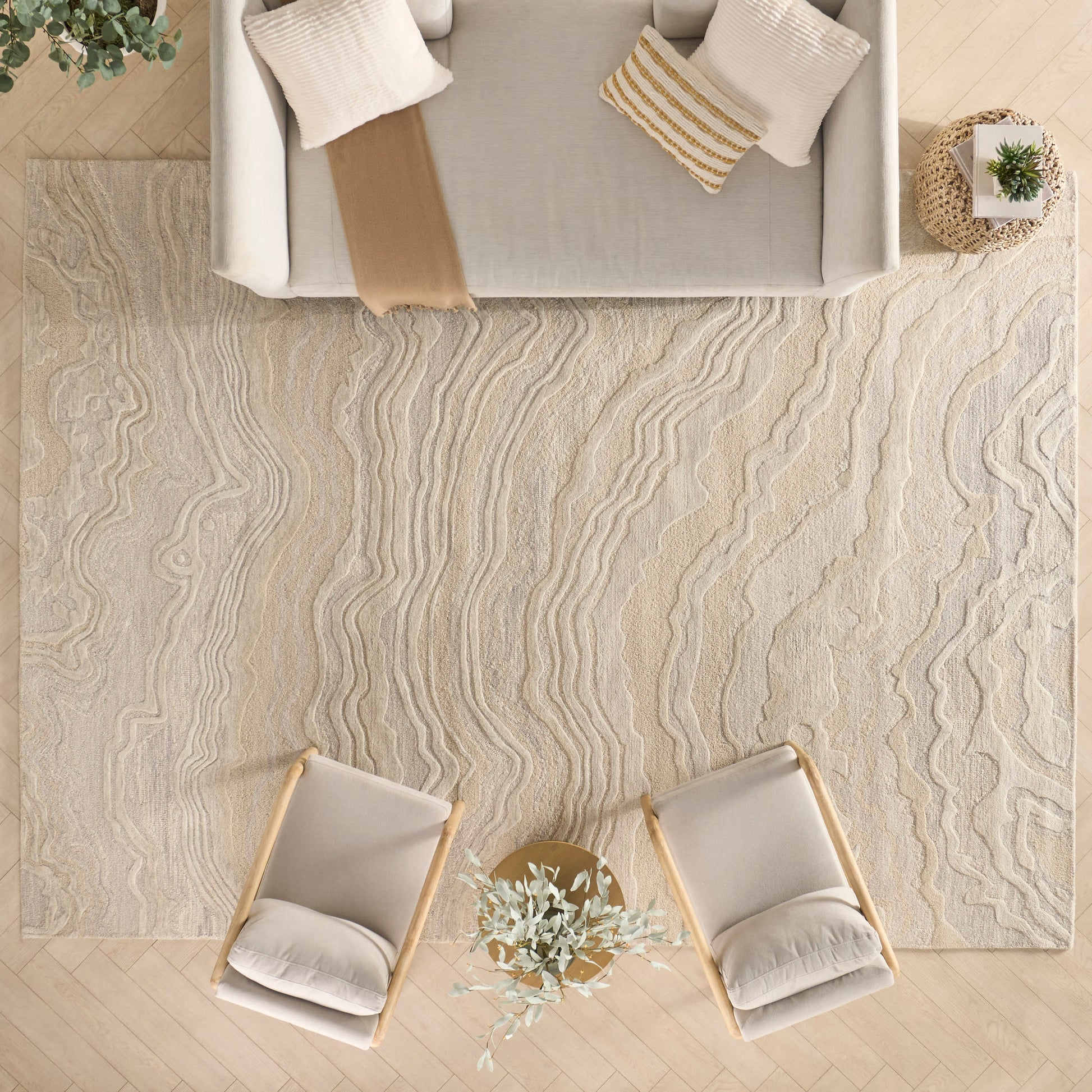 Top-down view: beige wavy rug, two cream armchairs around a small round table with a plant, beige sofa with cushions.