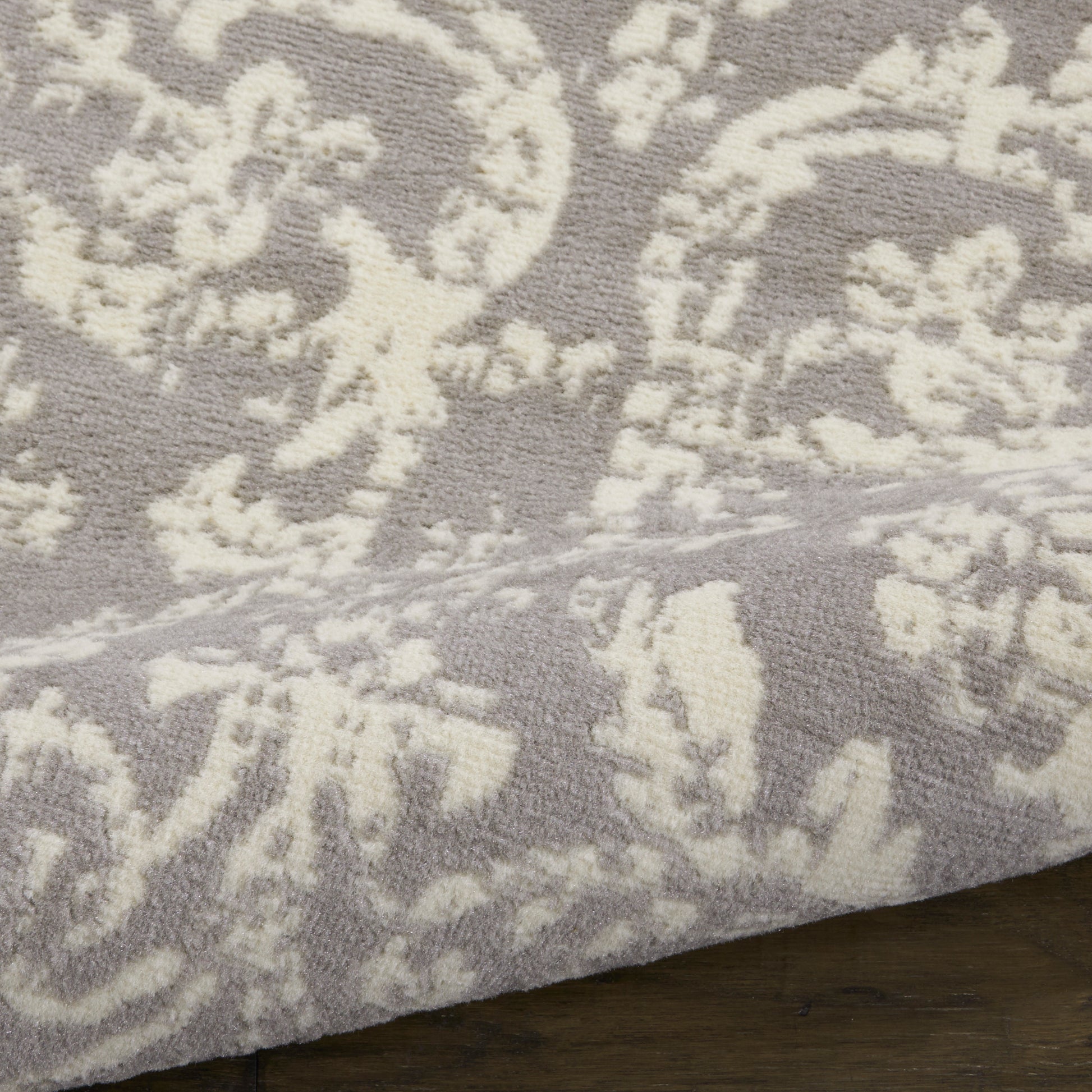 Lavender-gray plush rug with ivory damask floral pattern; soft tufted pile and visible edge on dark wood floor.
