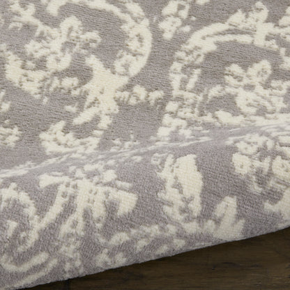 Lavender-gray plush rug with ivory damask floral pattern; soft tufted pile and visible edge on dark wood floor.