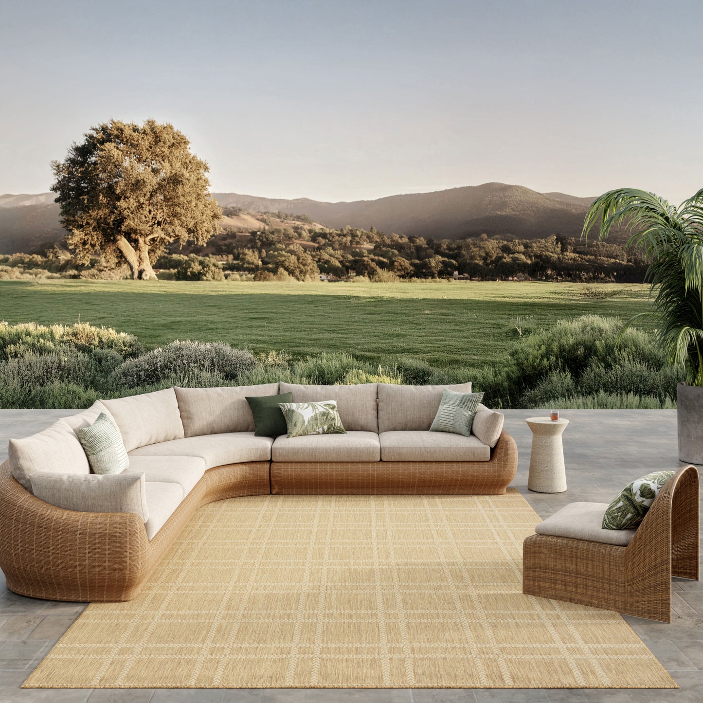 Outdoor tan rug with beige cushions and green pillows; scenic countryside backdrop.