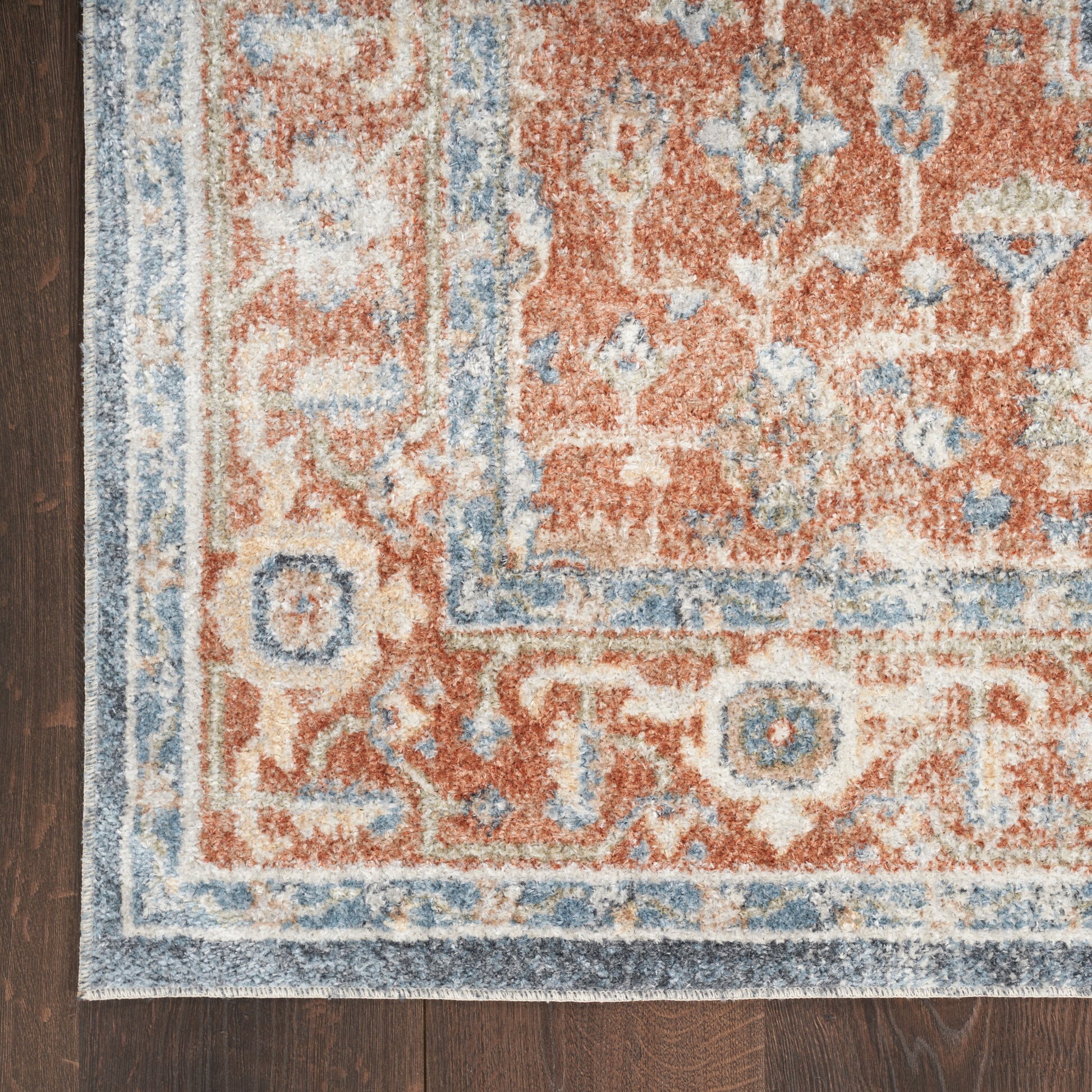 Close-up of a woven wool rug with red, blue and cream tones and a floral border on a dark wood floor.