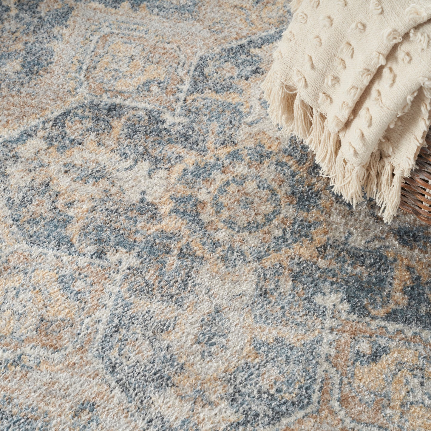Blue and beige ornate patterned area rug with a plush pile; cream fringed throw blanket at the corner.