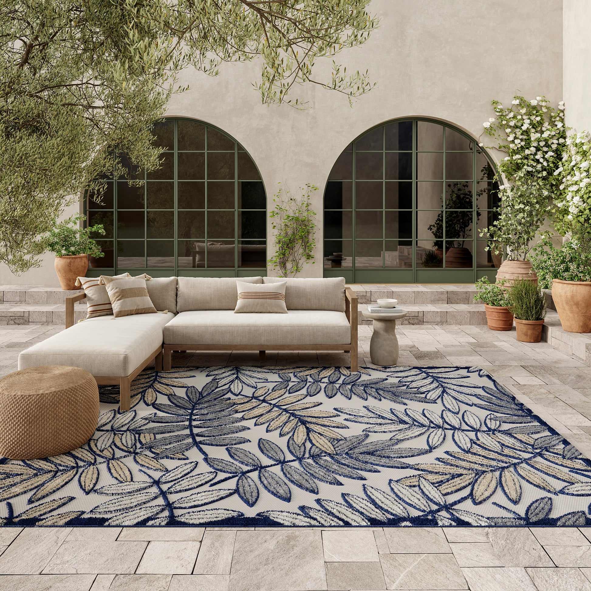 Outdoor patio with a beige sectional sofa, blue leaf-pattern rug, arched windows, and potted plants.