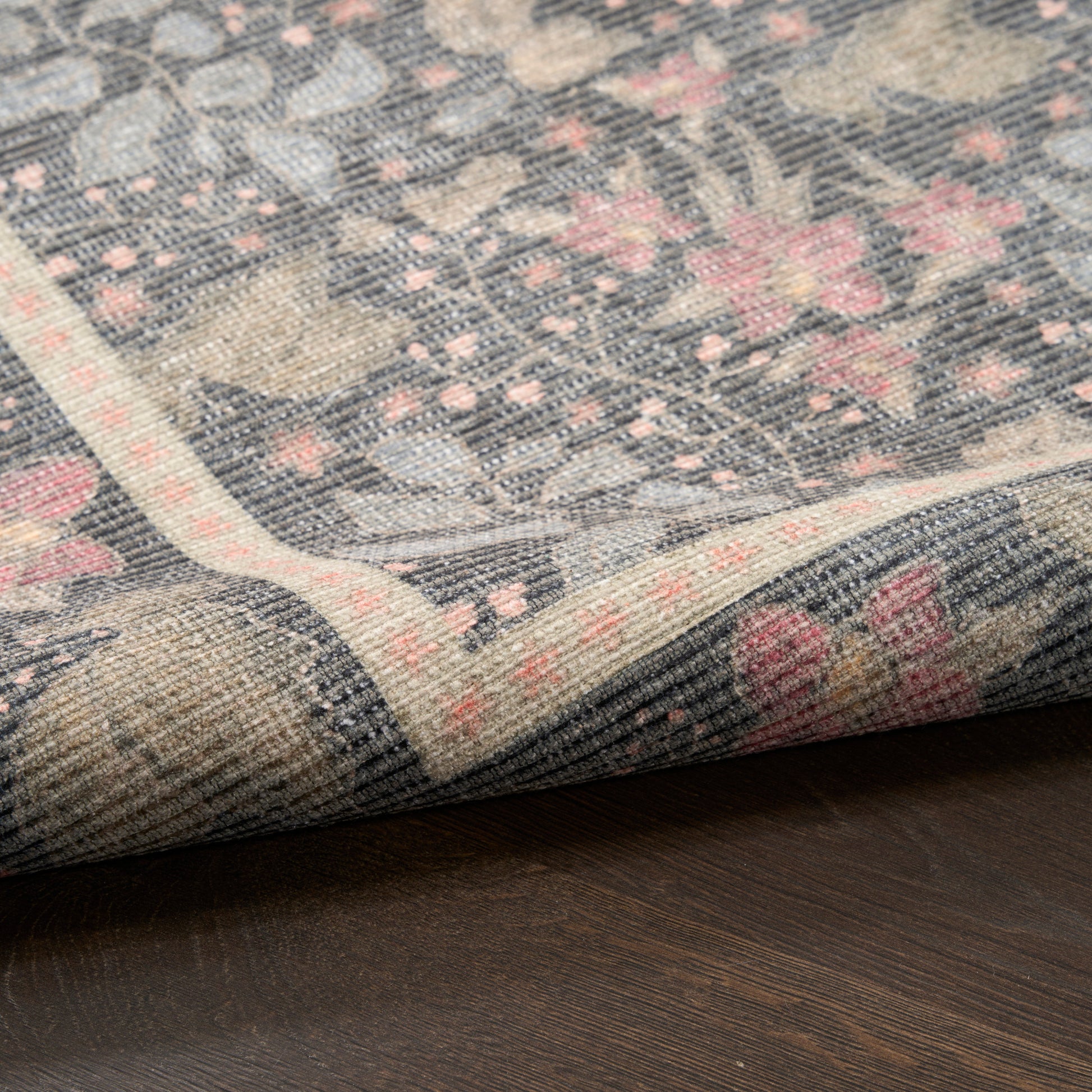 Blue-gray floral woven rug with pink blossoms and ivory border, folded corner on dark wood floor.
