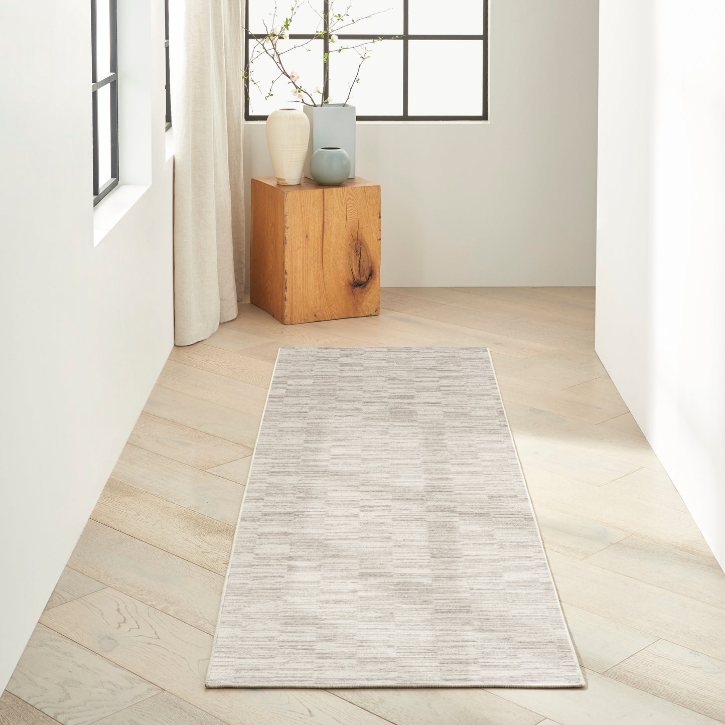 Light gray woven runner on pale wood flooring in a bright minimalist hallway, with a wooden block pedestal and ceramic vases.