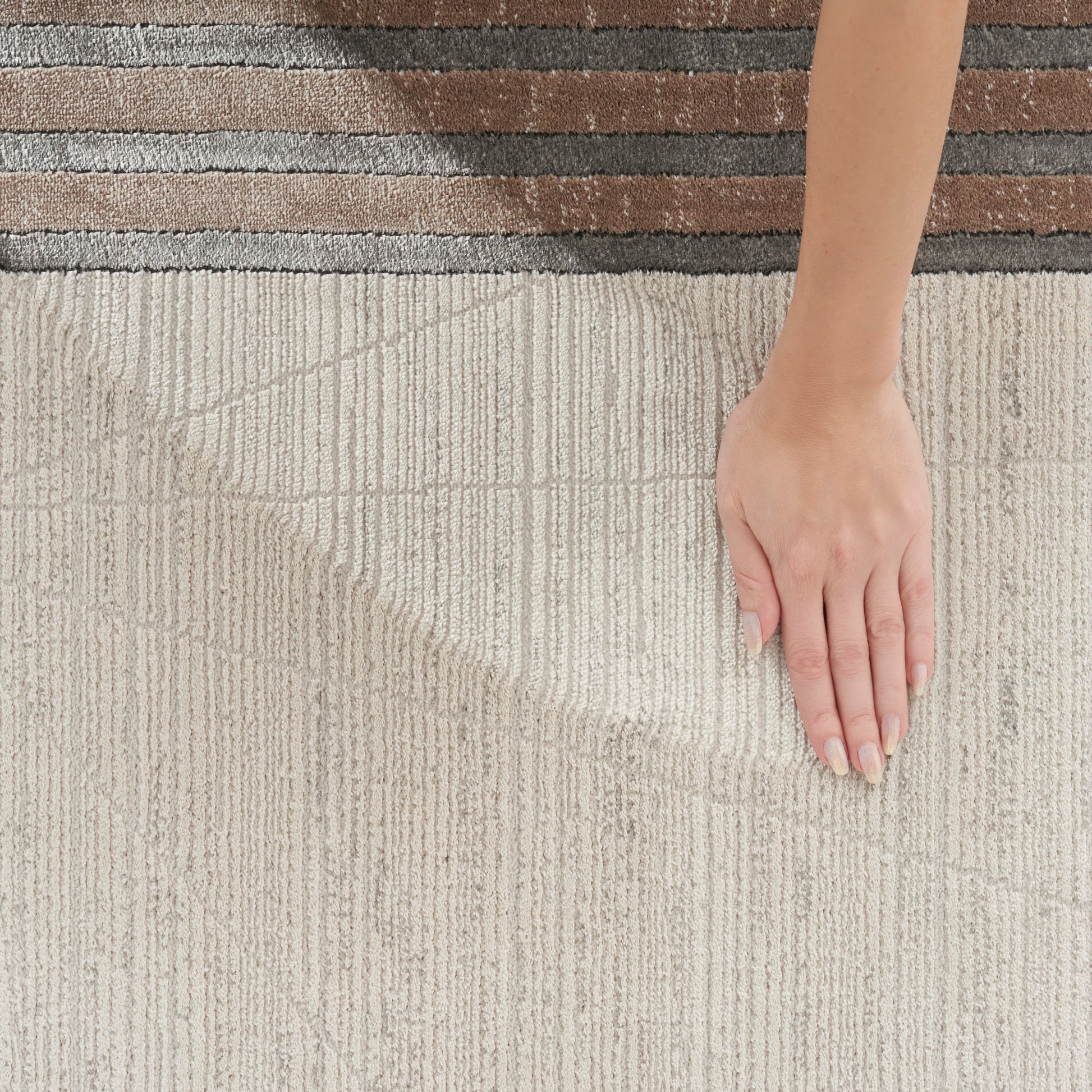 Beige textured runner rug with vertical ribs and brown border; a hand rests on the fabric, showing manicured nails.