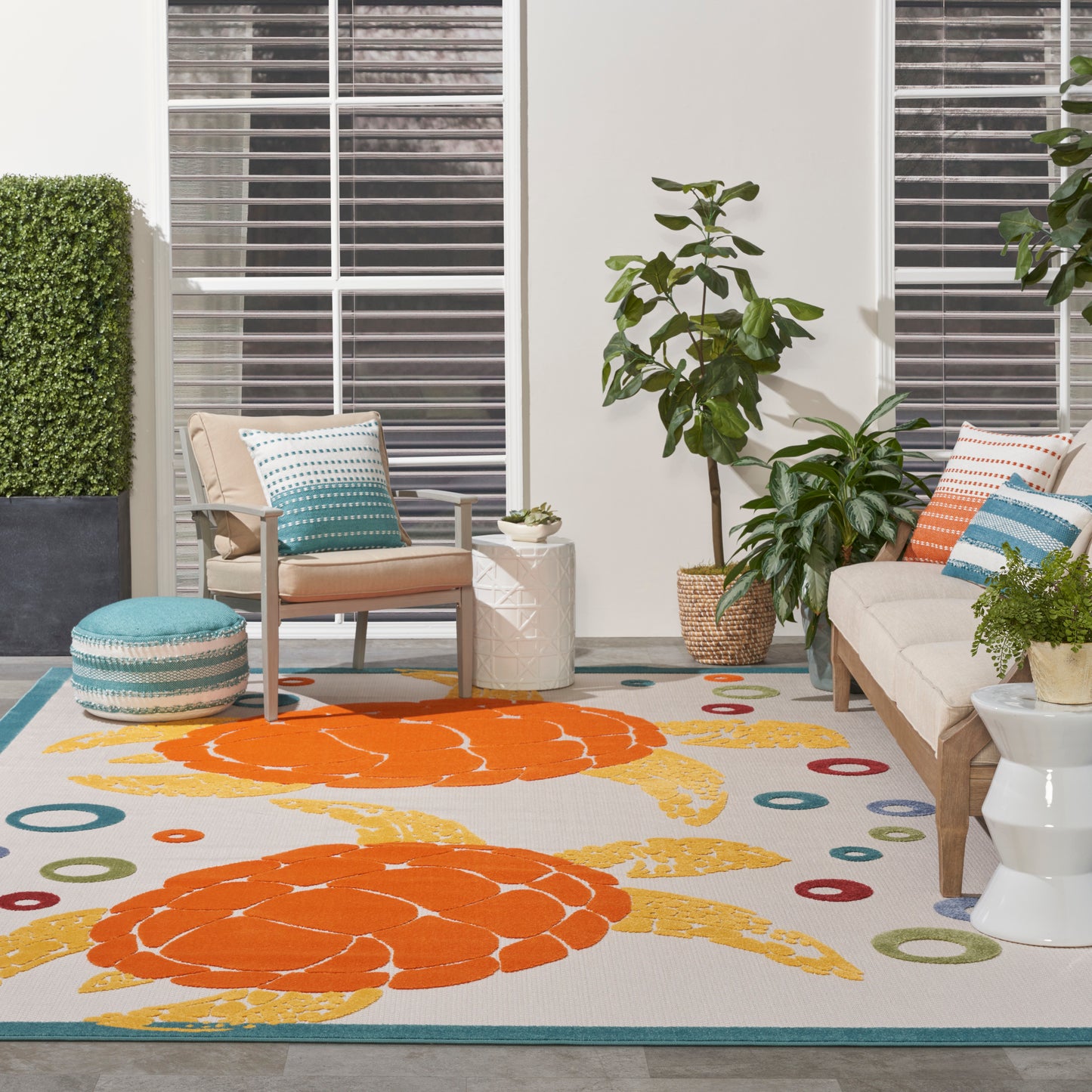 Outdoor patio with tan seating, teal cushions, round teal ottoman, vibrant rug with orange circles, & potted plants.